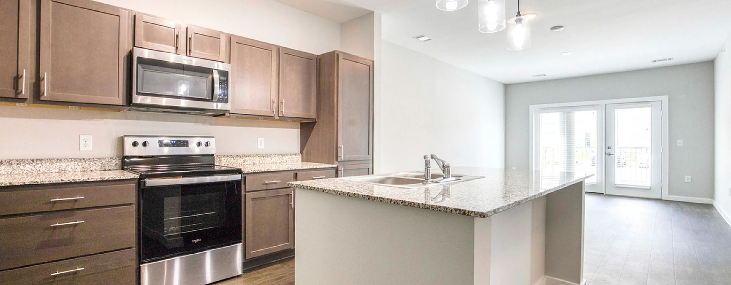Modern kitchen with granite countertops, sleek cabinetry, stainless steel appliances, and an open layout leading to a bright living area with glass doors.