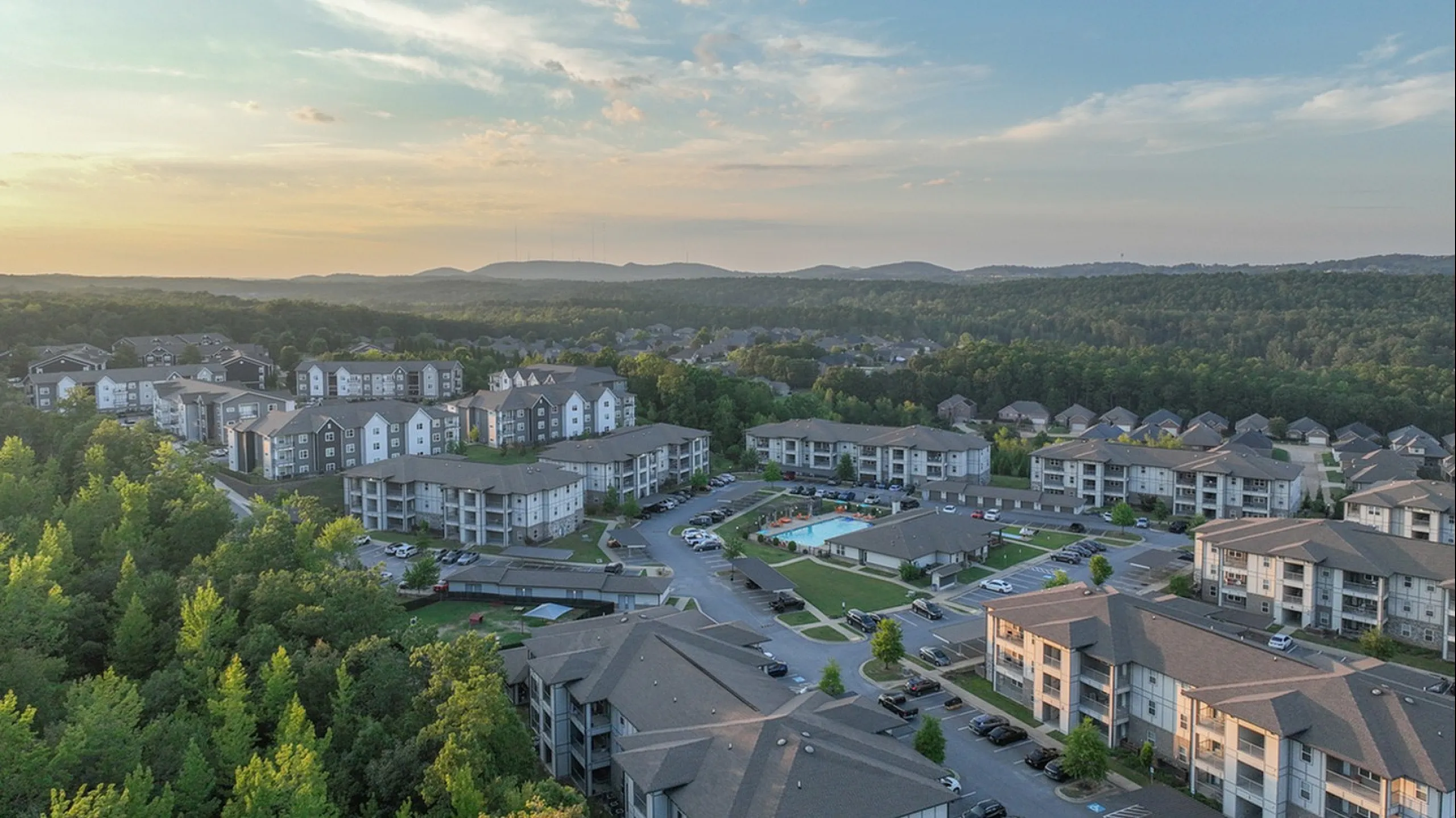 Scenic aerial view of a vibrant apartment community surrounded by lush greenery, offering modern living spaces and a relaxing environment at sunset.