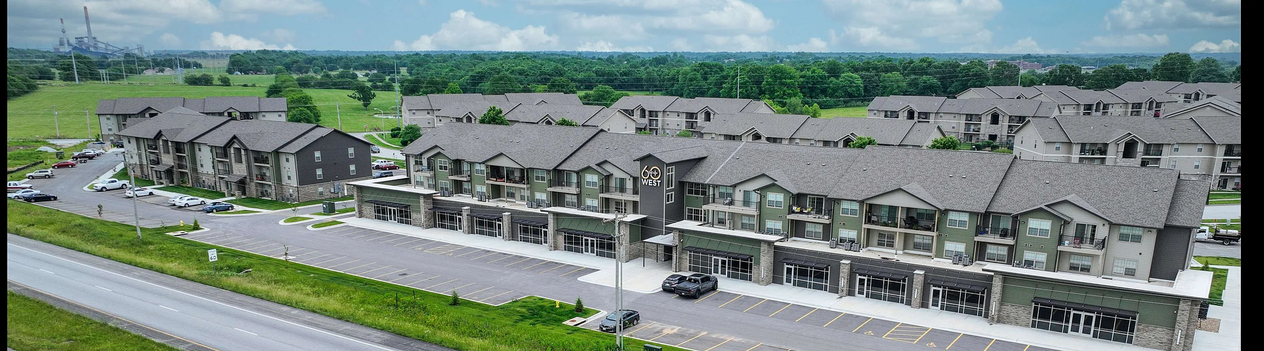 Modern apartment complex with sleek design nestled in a lush green setting, offering ample parking and convenient highway access.