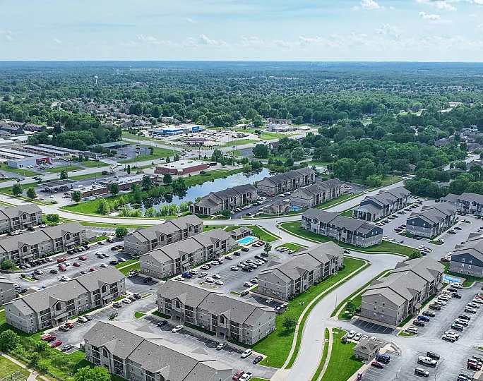Expansive aerial view of a vibrant residential community featuring modern apartment buildings, scenic green spaces, and a central pool area, surrounded by lush greenery.