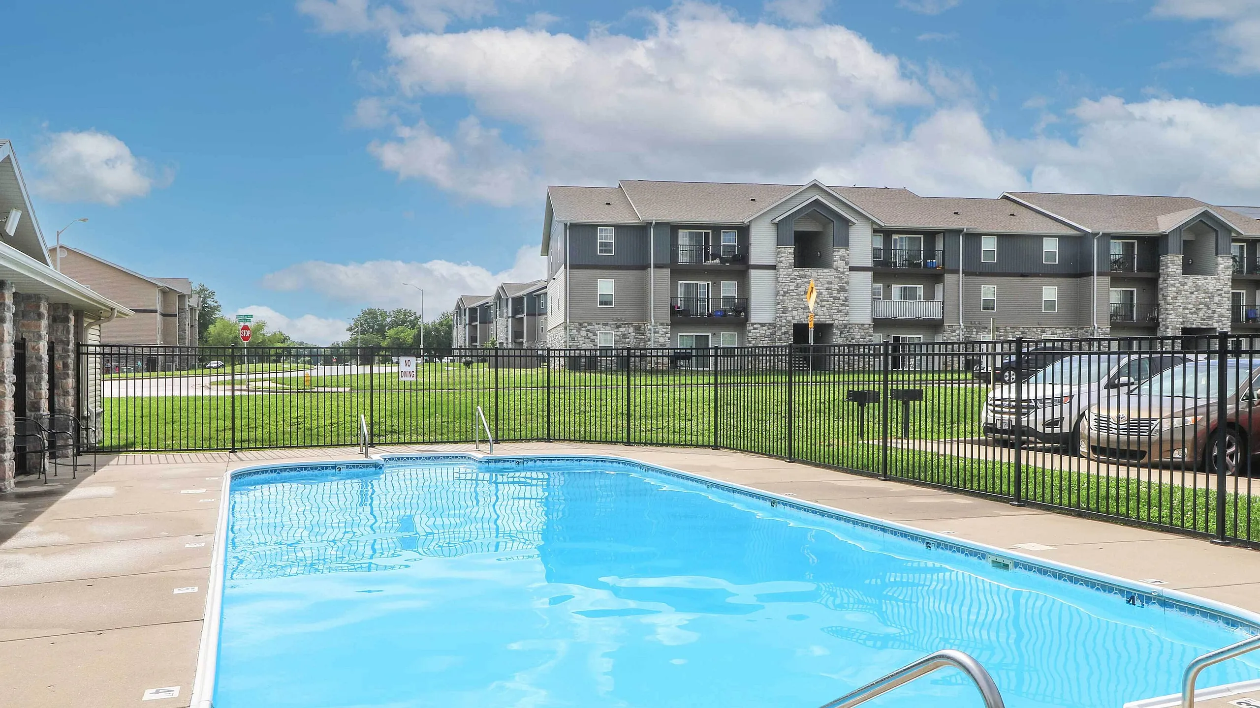Inviting apartment complex with a sparkling outdoor pool, modern architecture, and lush green surroundings under a clear blue sky.