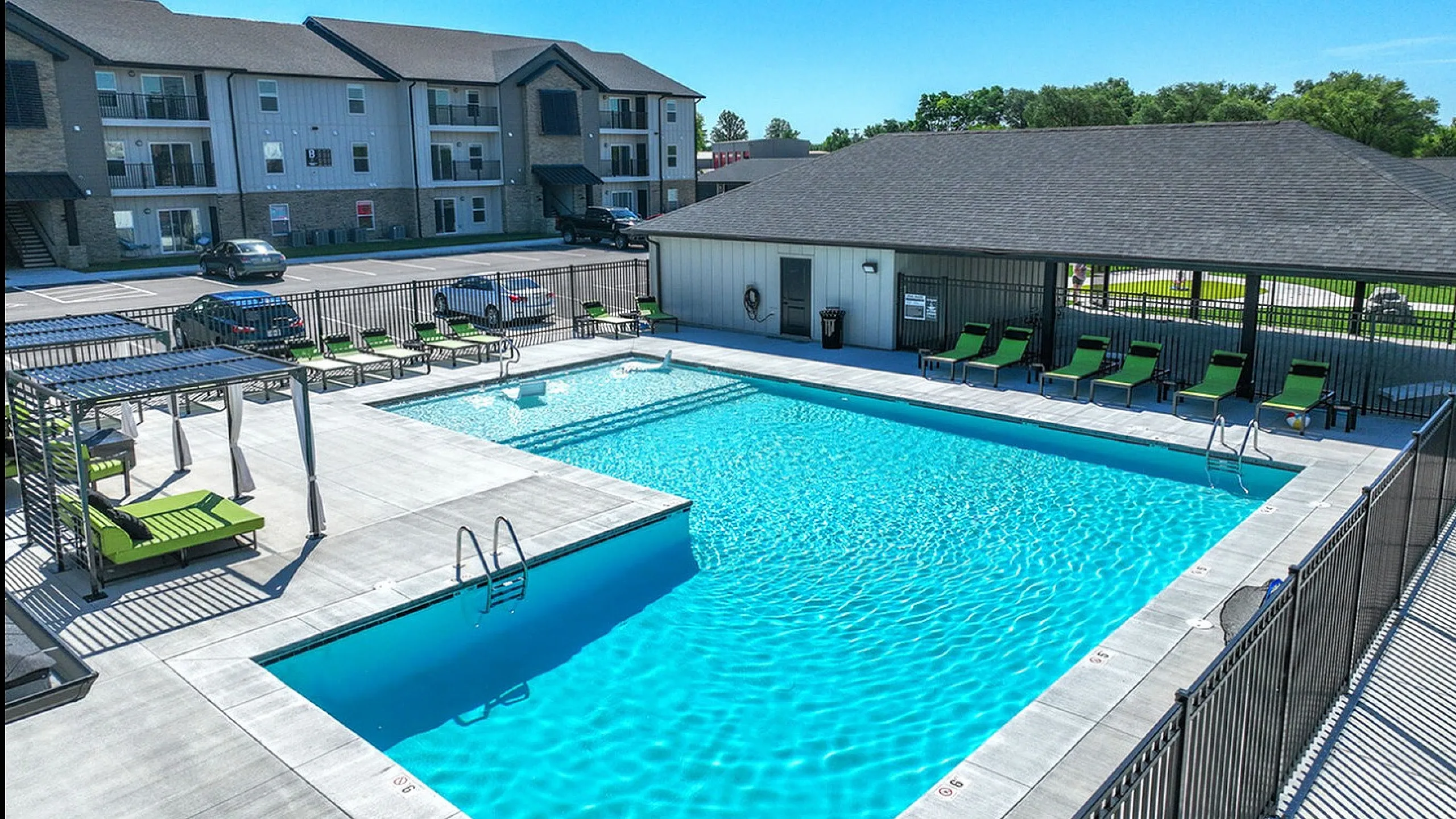 Inviting apartment complex featuring a sparkling blue swimming pool surrounded by sleek green lounge chairs and pergola seating, perfect for relaxation and leisure.