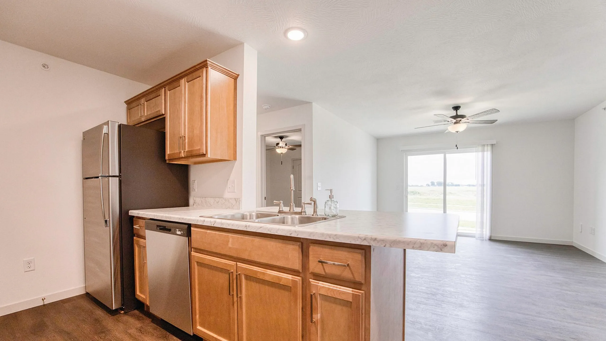 Bright and modern kitchen with wooden cabinetry and sleek stainless steel appliances, opening into a spacious living area with a ceiling fan and large sliding glass doors.
