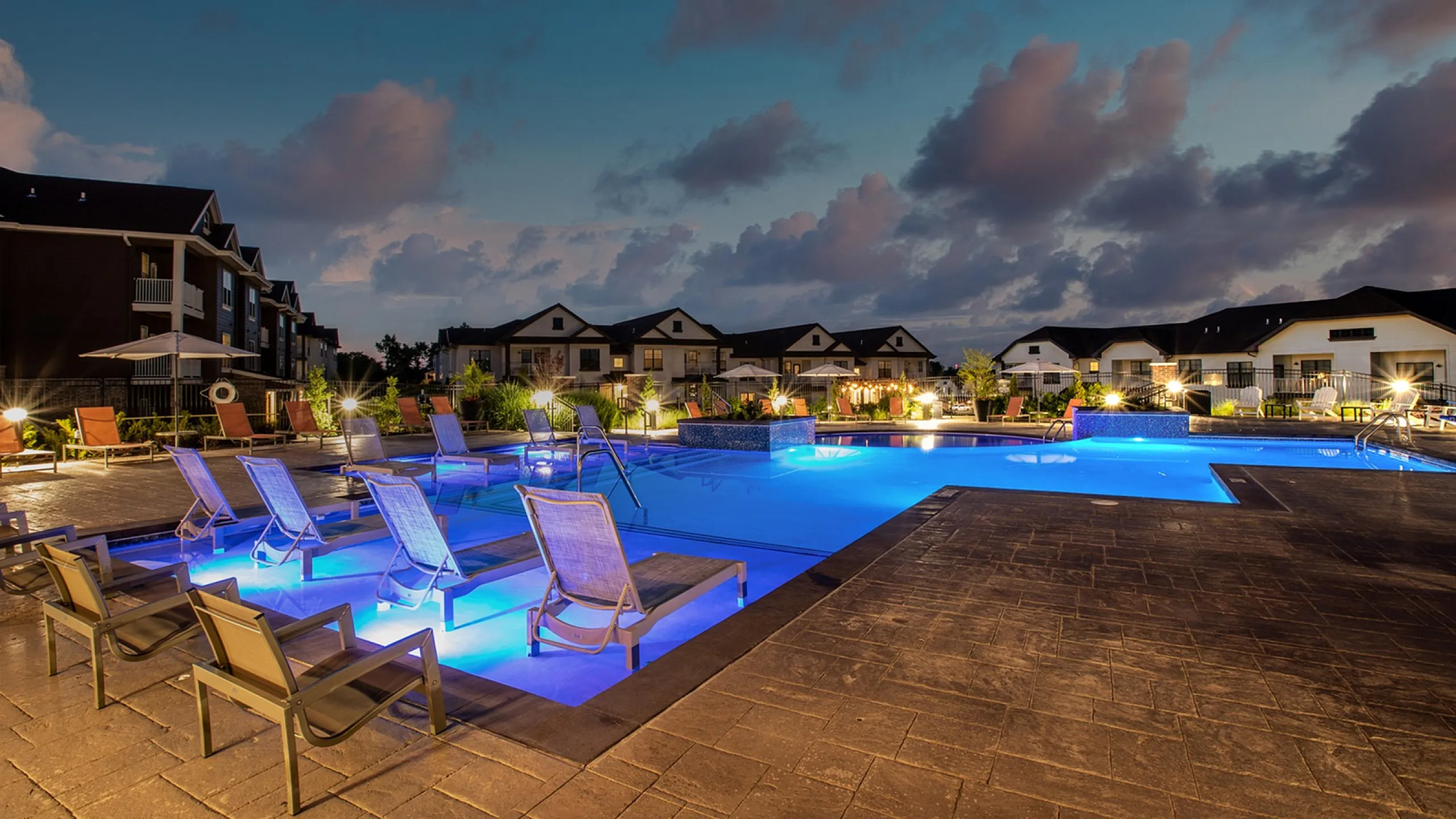 Luxurious apartment with stunning evening pool area, ambient lighting, and chic lounge chairs under a vibrant dusk sky.