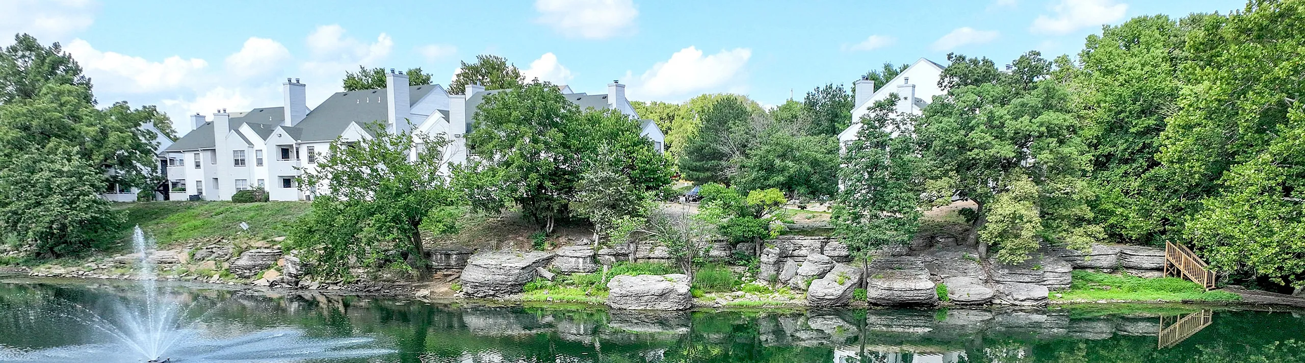 Picturesque lakeside view of elegant white apartment buildings surrounded by lush greenery, featuring a serene fountain reflecting in the tranquil water.