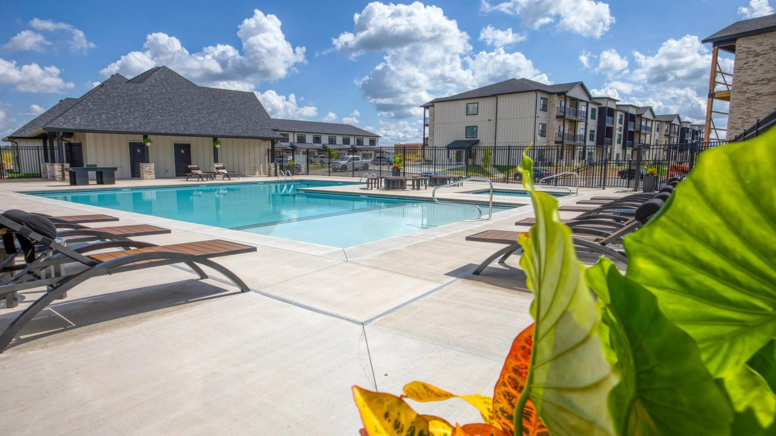 Chic apartment community with a pristine swimming pool, sleek lounge chairs, and vibrant landscaping under a bright blue sky.