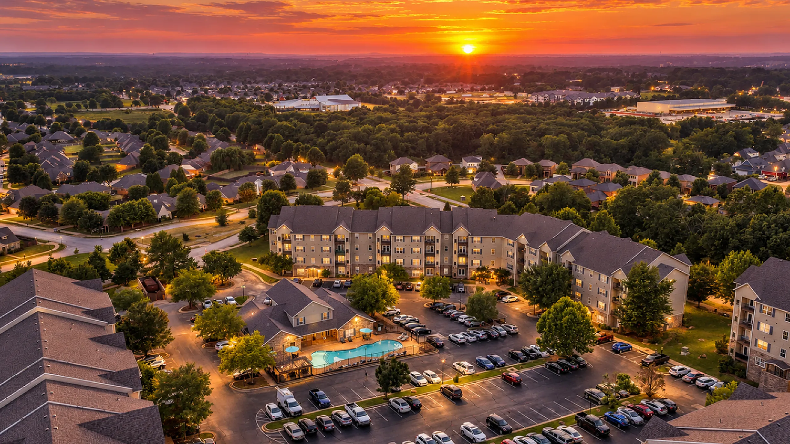 Charming aerial view of a vibrant apartment complex surrounded by lush greenery at sunset, showcasing a central communal pool and ample parking.