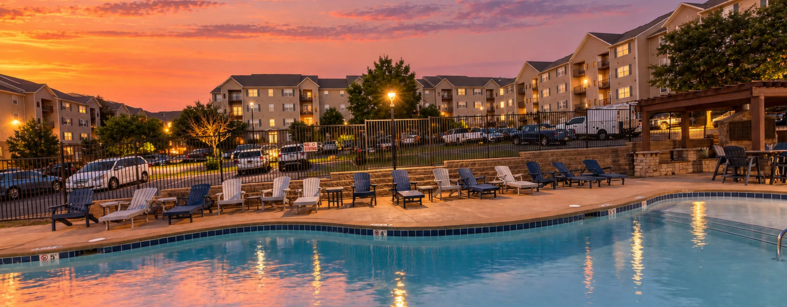 Charming apartment community featuring a beautifully lit pool area at sunset, surrounded by inviting lounge chairs and picturesque residential buildings.
