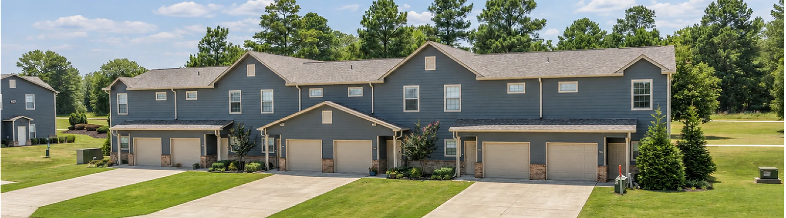 Charming townhouse complex with modern gray exterior, private garages, and lush green lawns, set against a backdrop of serene natural surroundings.
