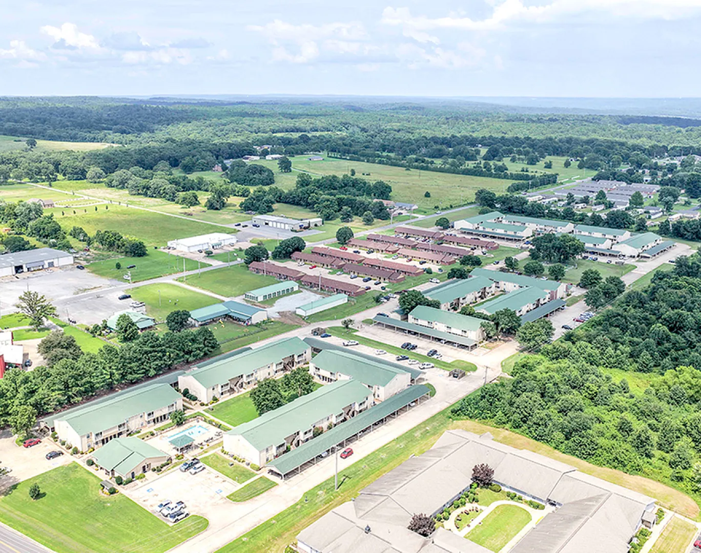 Aerial view of a charming residential complex surrounded by lush greenery, featuring modern apartments with green rooftops and spacious parking areas.