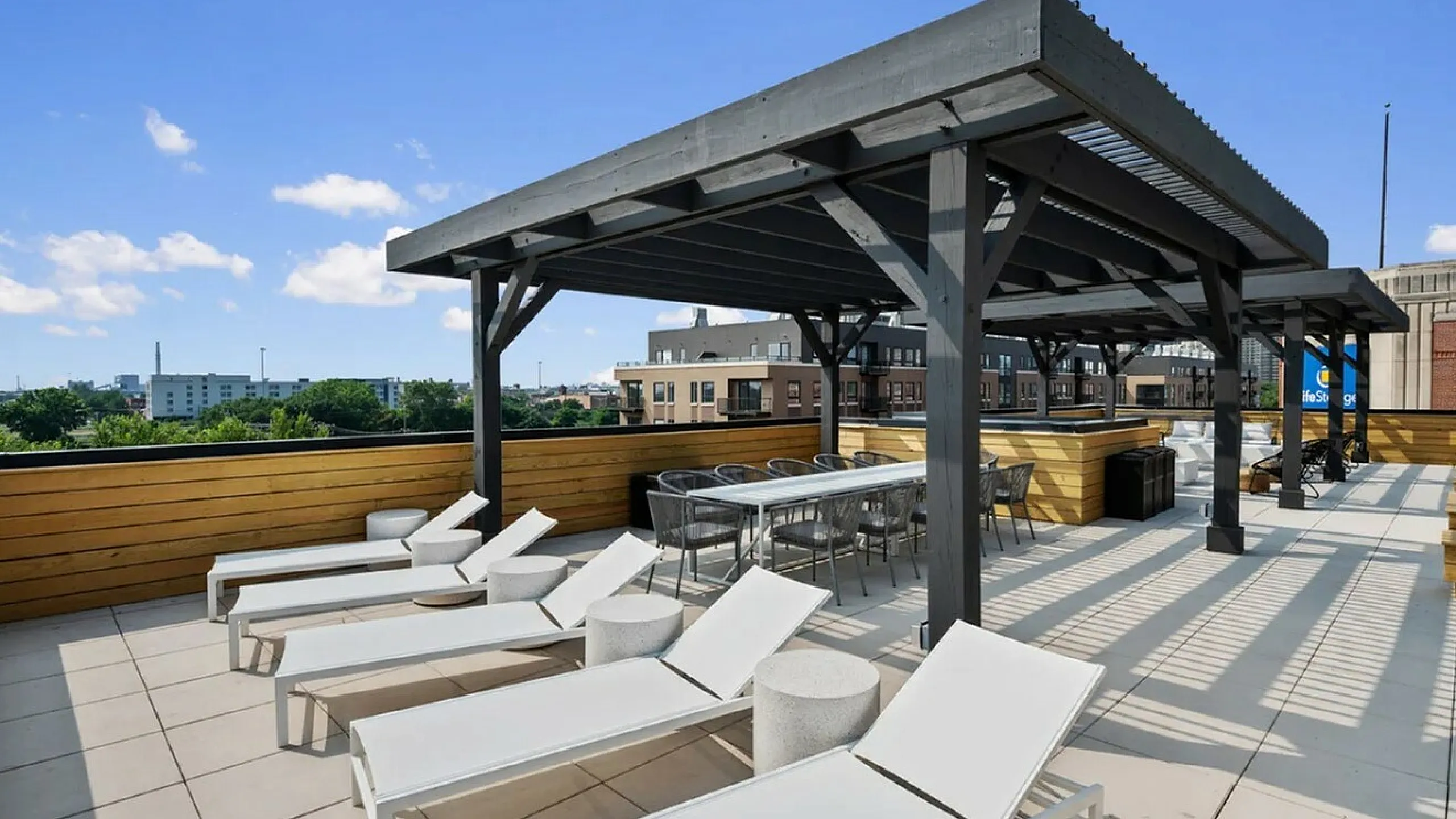 Chic urban rooftop terrace with elegant sun loungers and a shaded dining area, perfect for relaxing or entertaining under a bright blue sky.