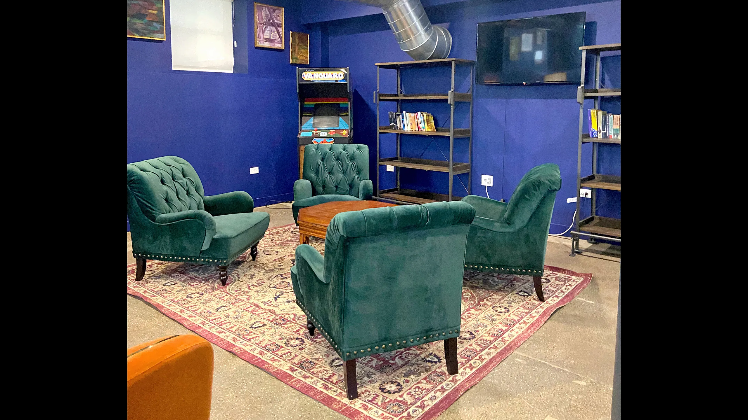 Chic lounge area with emerald green velvet chairs, vintage arcade game, and elegant bookshelves in a stylish blue room.