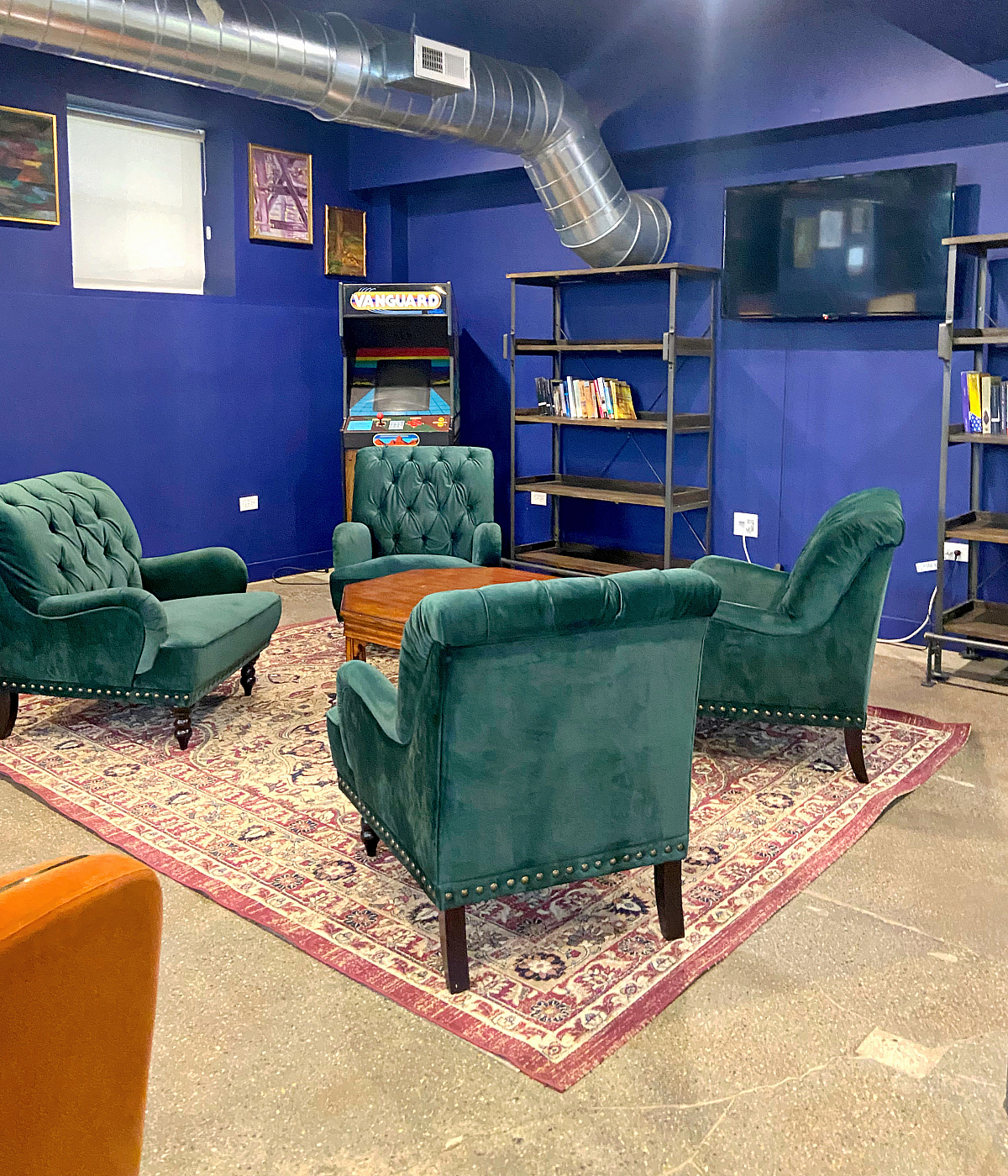 Chic lounge area with plush green armchairs, stylish rug, and vibrant blue walls featuring modern artwork and a vintage arcade machine.