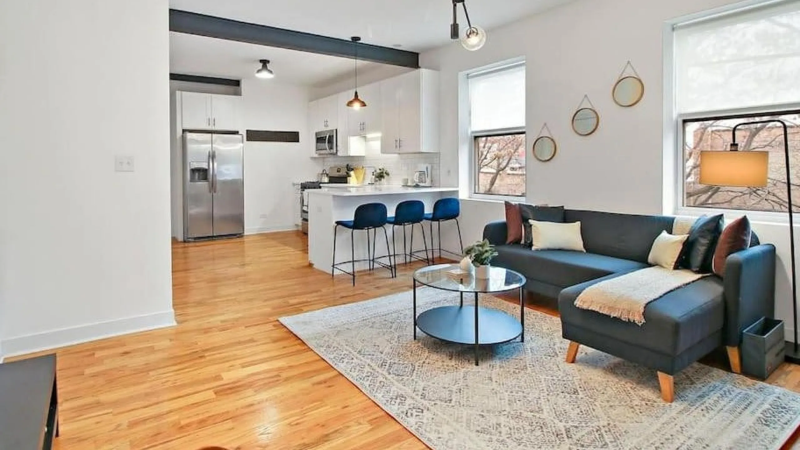 Inviting open-plan living room with chic decor, cozy sectional sofa, modern light fixture, and sleek kitchen with stainless steel appliances.
