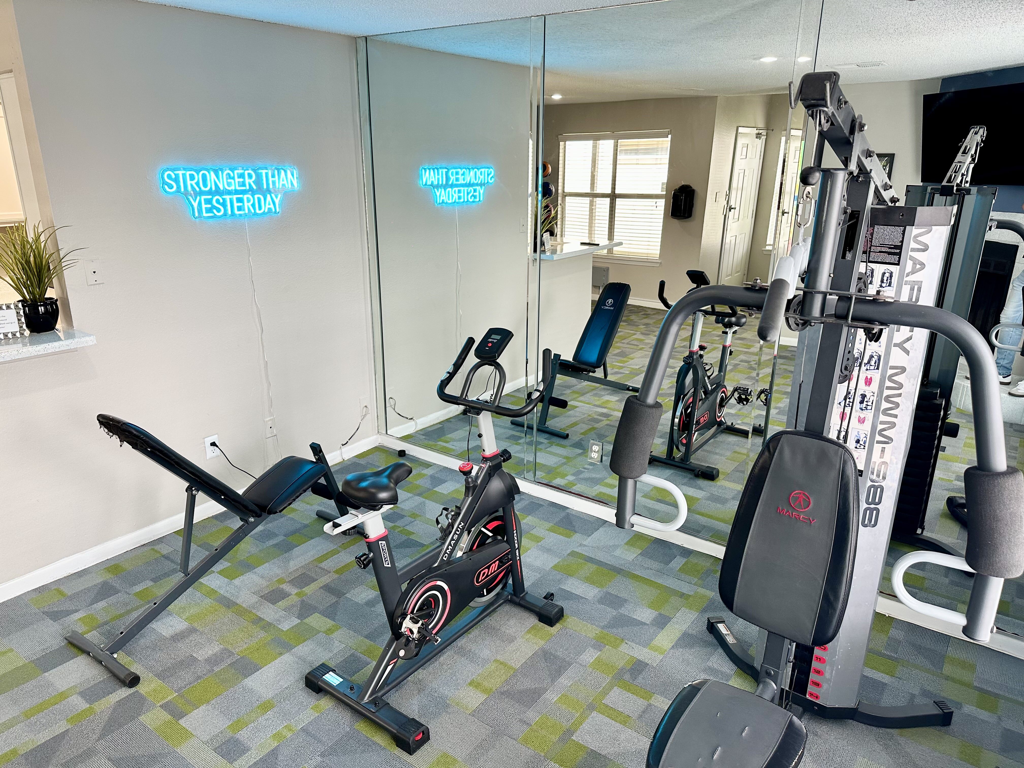 Modern apartment gym featuring a stationary bike, weight bench, and full-length mirrors, illuminated by motivational neon lighting.