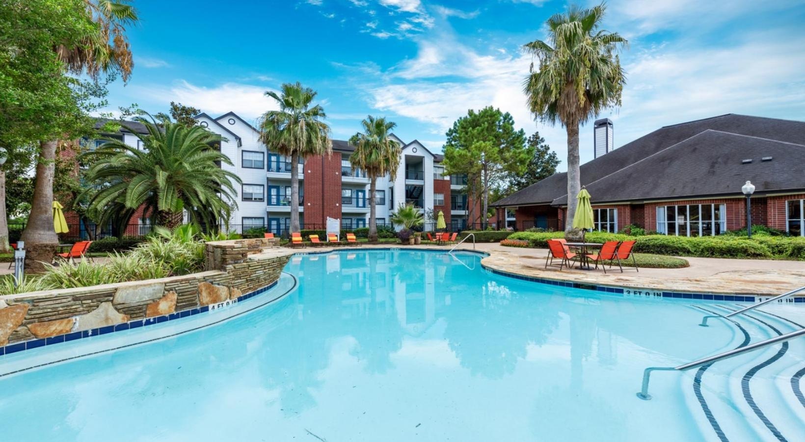Luxurious apartment complex with a pristine outdoor pool, lush palm trees, and stylish brick architecture under a vibrant blue sky.