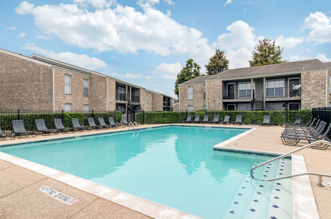 Inviting apartment complex featuring a pristine swimming pool surrounded by comfortable lounge chairs, set against a backdrop of modern two-story buildings and a clear blue sky.