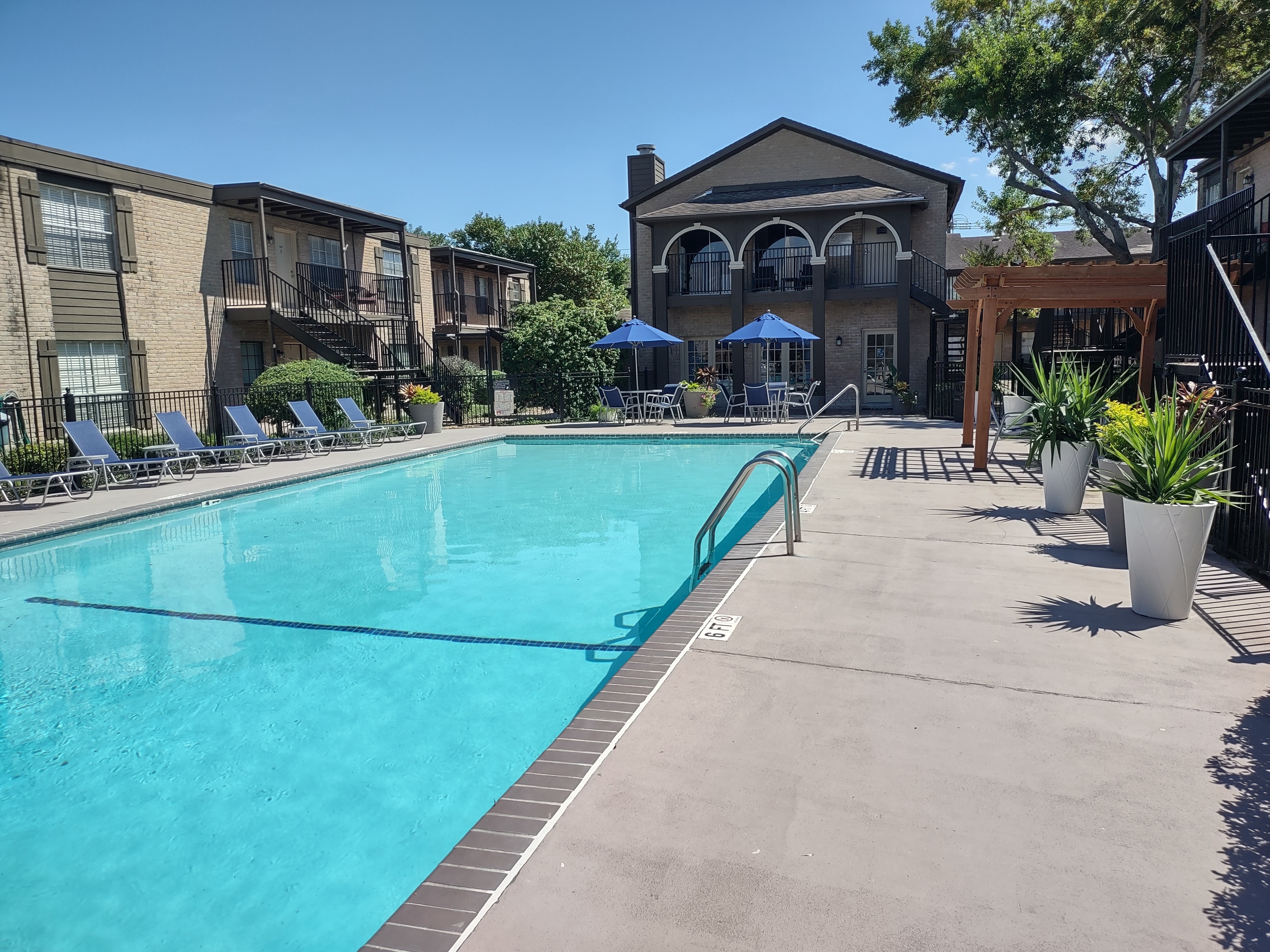Inviting apartment pool area with sun loungers, shady umbrellas, and lush greenery on a bright day.
