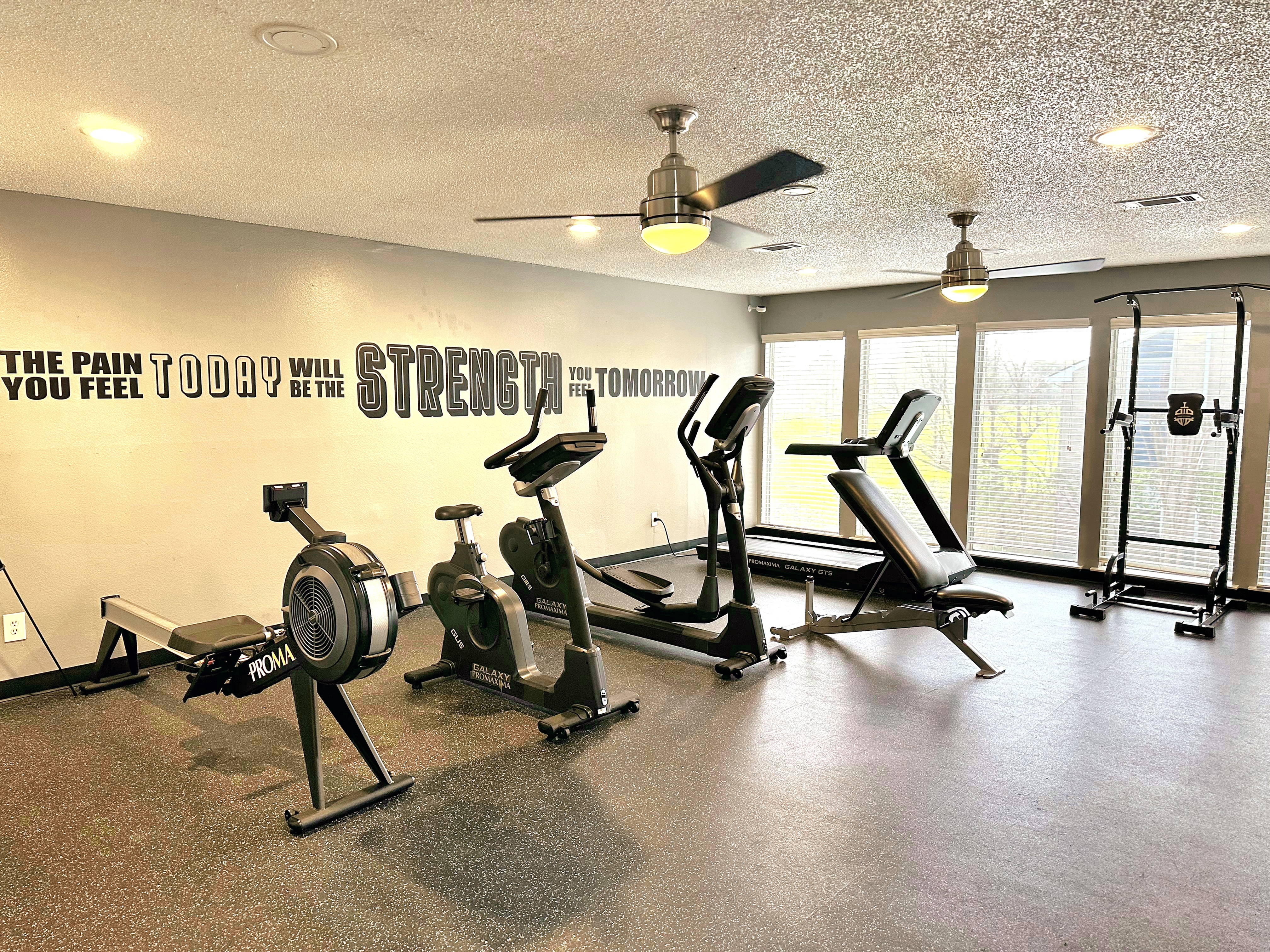 Modern apartment gym featuring state-of-the-art exercise equipment, motivational wall quote, and bright natural lighting from large windows.