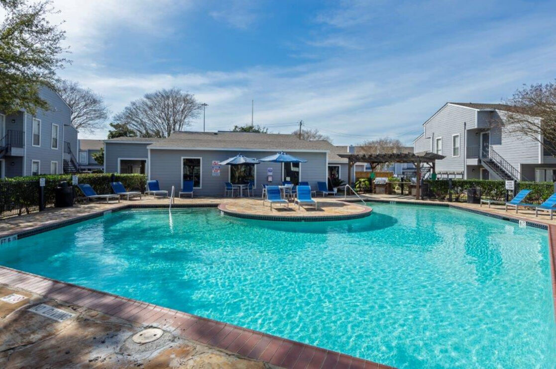 Inviting outdoor pool area with sun loungers and blue umbrellas, surrounded by modern apartments and lush greenery.