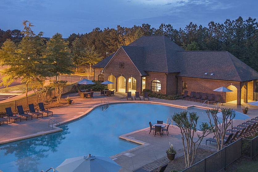 Charming evening view of a luxurious apartment complex with a sparkling outdoor pool, stylish lounge chairs, and elegant brick clubhouse surrounded by lush greenery.