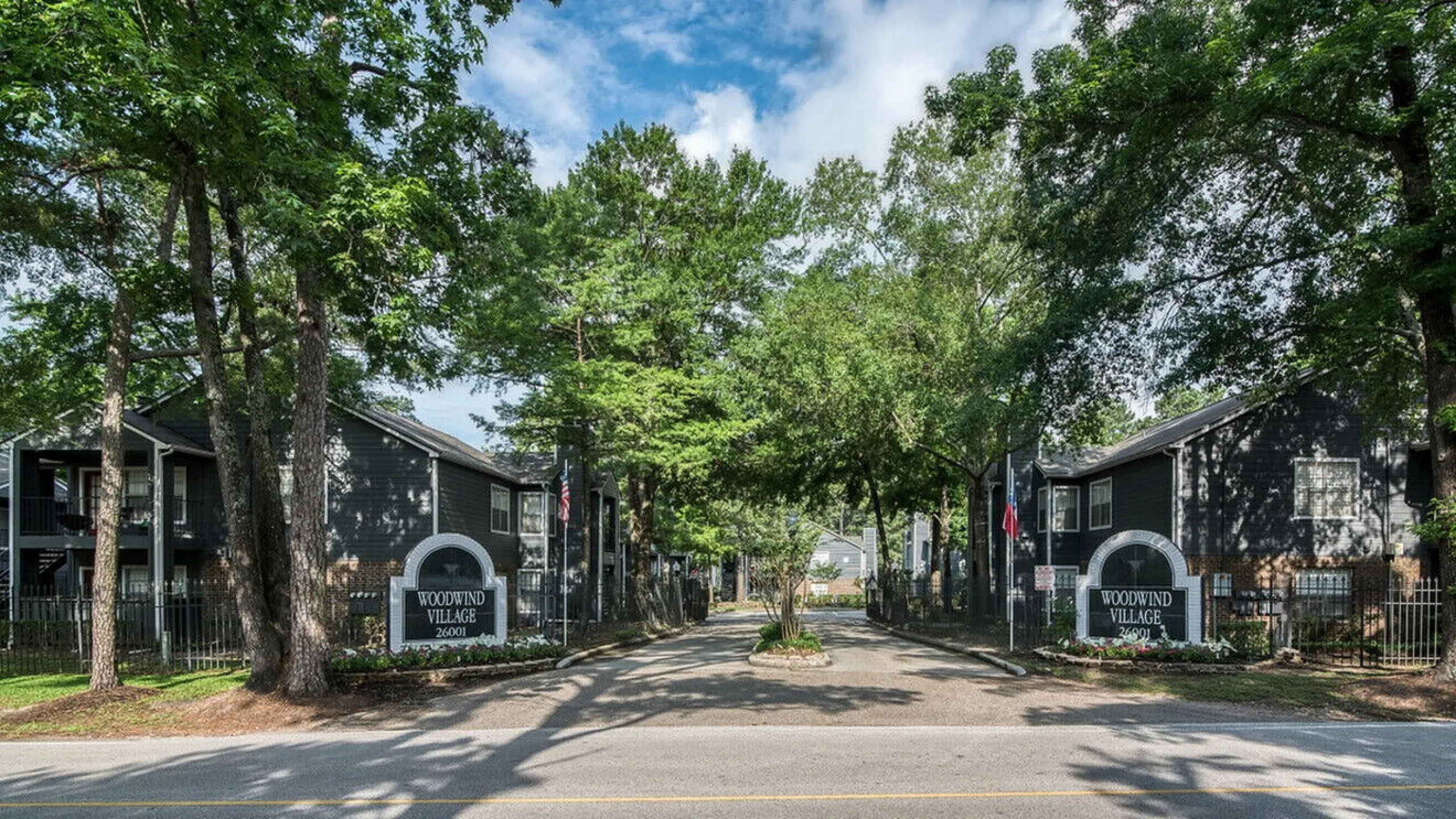 Charming entrance to Woodwind Village, featuring lush trees and welcoming signage set against elegant residential buildings.