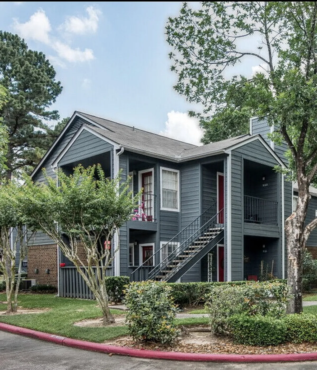 Charming two-story apartment complex with stylish gray exteriors, surrounded by lush trees and ample parking, offering a serene and inviting living environment.