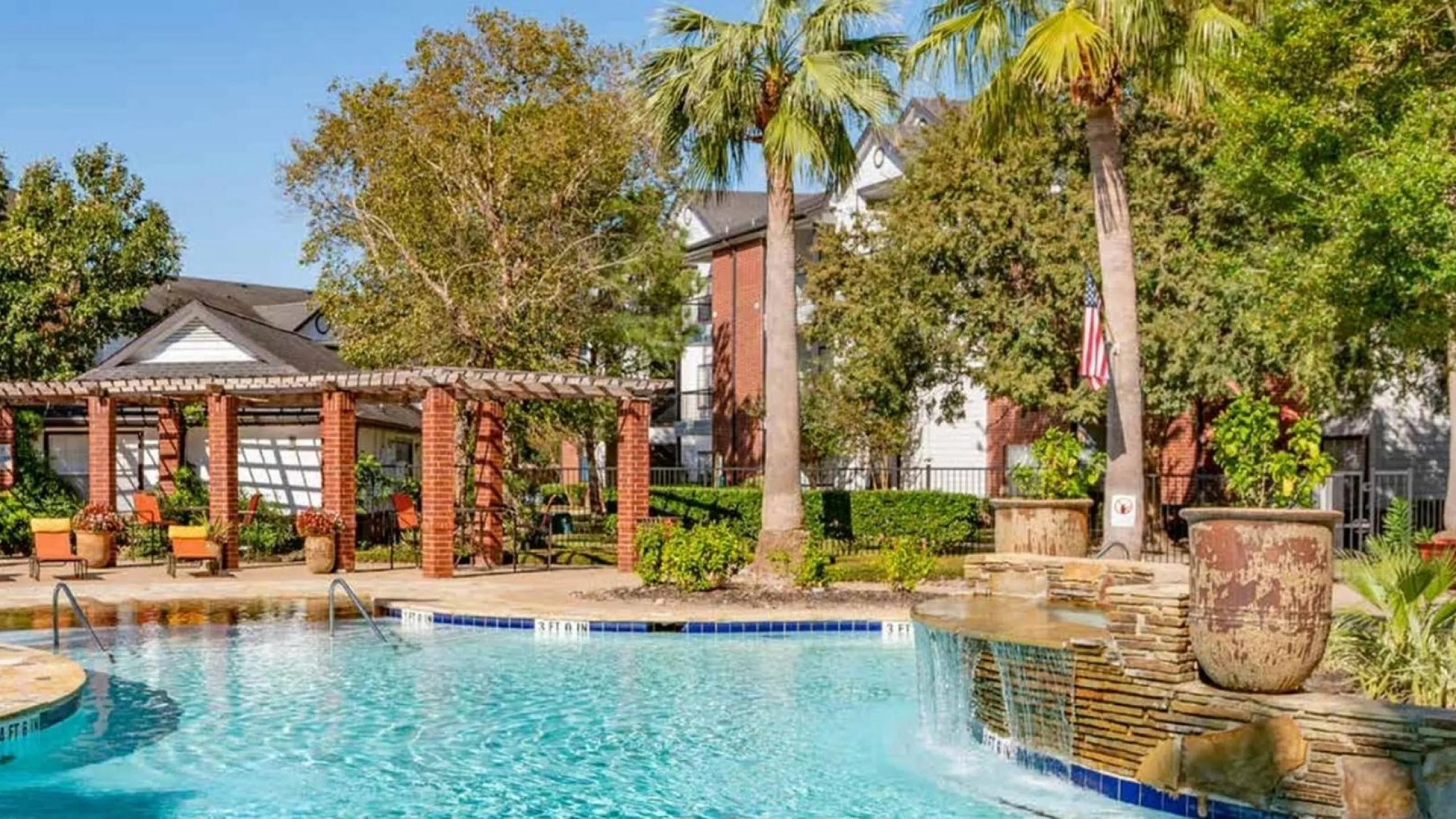 Lush apartment complex with a sparkling pool, surrounded by vibrant greenery and elegant brick pergolas, ideal for relaxation.
