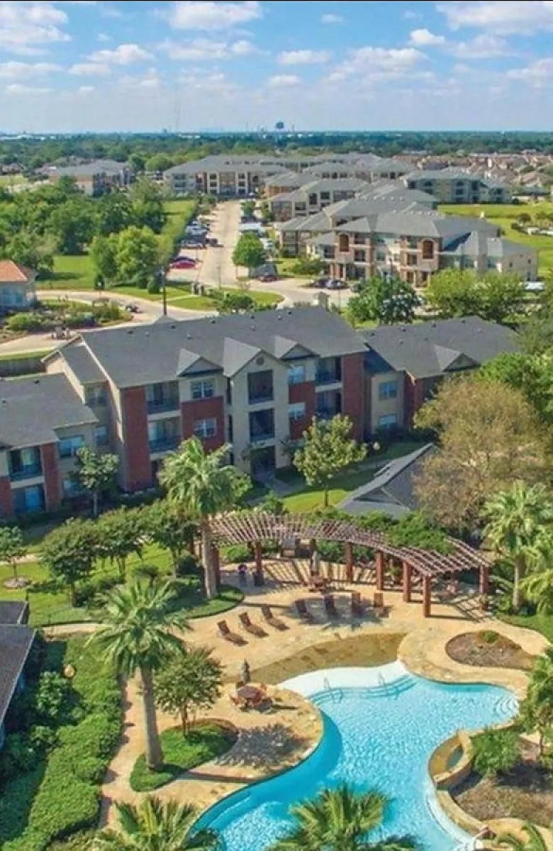 Aerial view of a vibrant apartment complex featuring lush landscaping, a resort-style pool, and modern buildings under a clear blue sky.