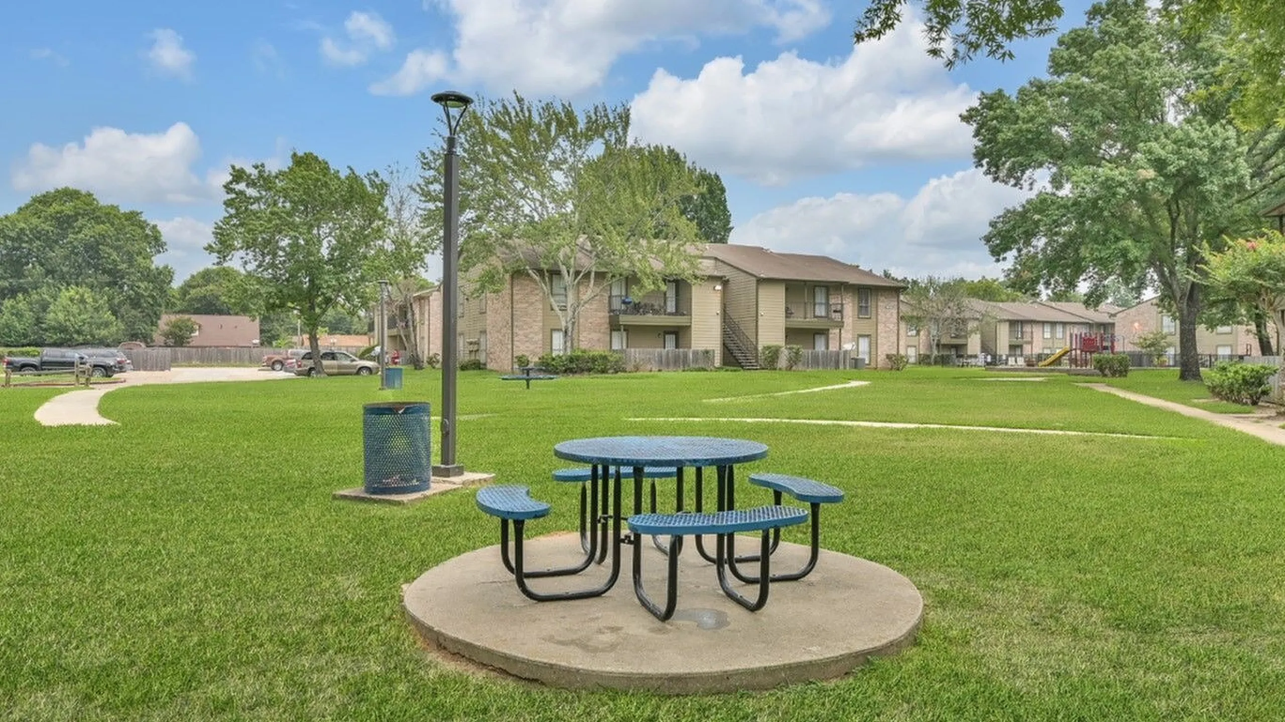 Inviting apartment complex with spacious green lawns, a cozy picnic area, and charming residential buildings under a vibrant blue sky.