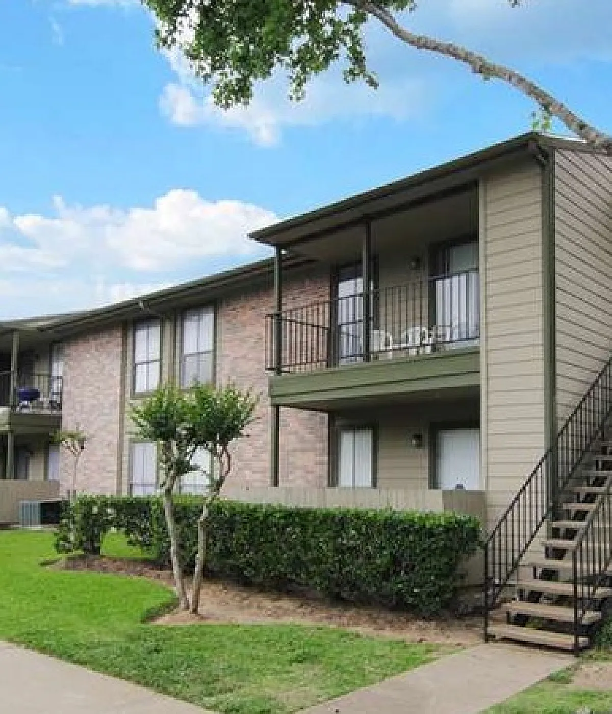 Charming apartment building with private balconies, lush greenery, and a welcoming pathway under a blue sky.