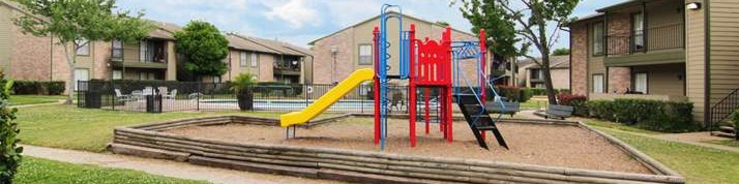 Charming apartment complex featuring a vibrant playground, lush green lawns, and cozy two-story buildings under a bright blue sky.