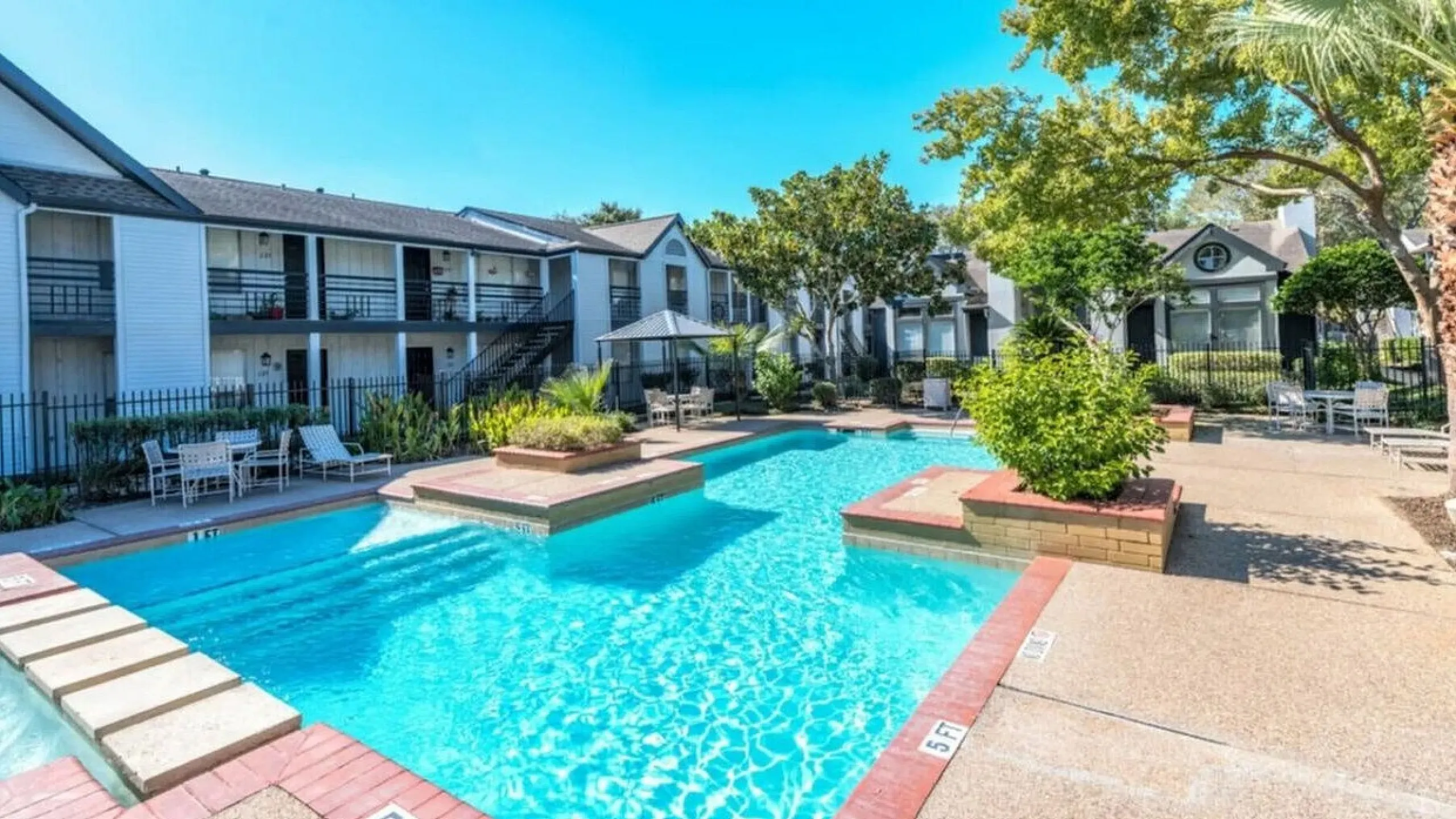 Inviting apartment complex featuring a sparkling swimming pool, surrounded by lush greenery and comfortable outdoor seating.
