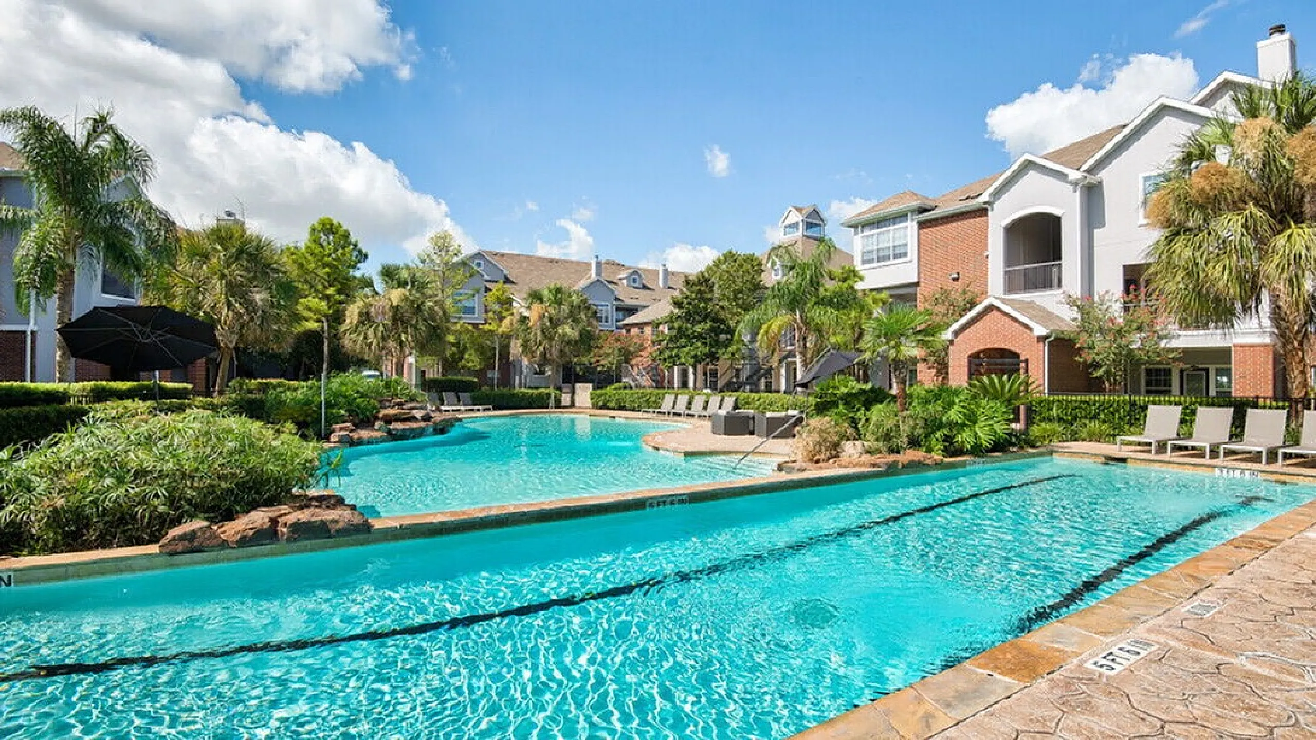 Luxurious apartment complex with a sparkling swimming pool, surrounded by lush palm trees and modern architecture under a bright blue sky.