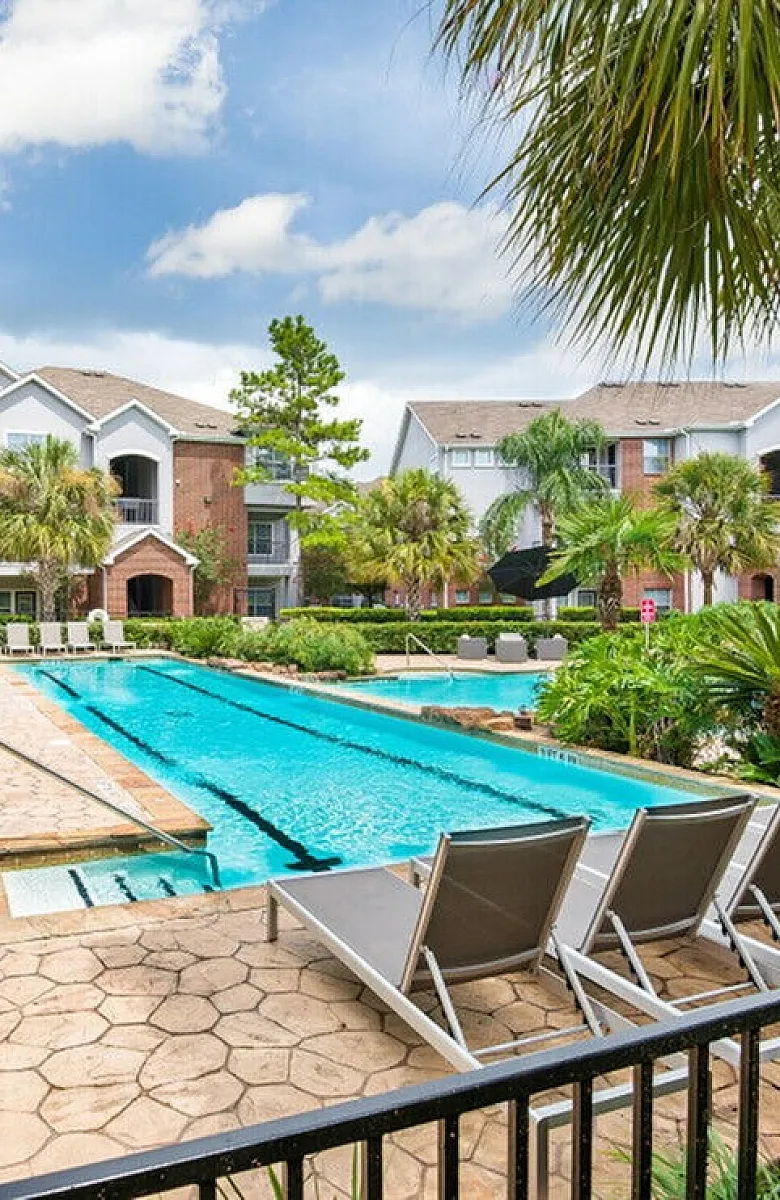 Luxurious apartment complex with a sparkling pool, surrounded by lush palm trees and comfortable lounge chairs under a clear blue sky.