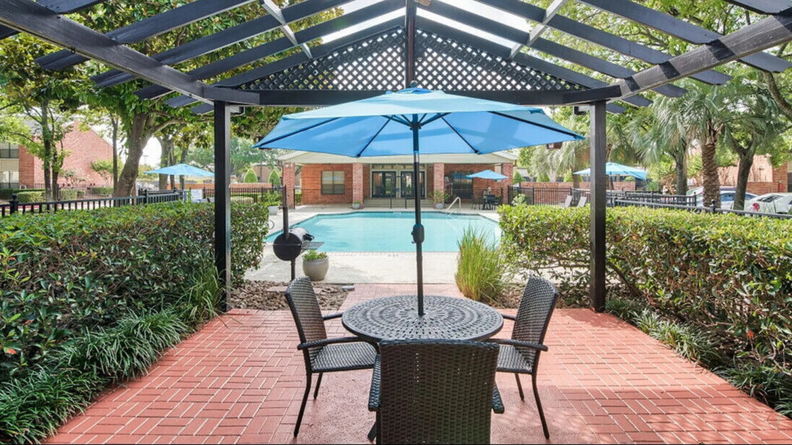 Inviting outdoor patio with a stylish seating area and vibrant blue umbrella, overlooking a sparkling pool surrounded by lush greenery.