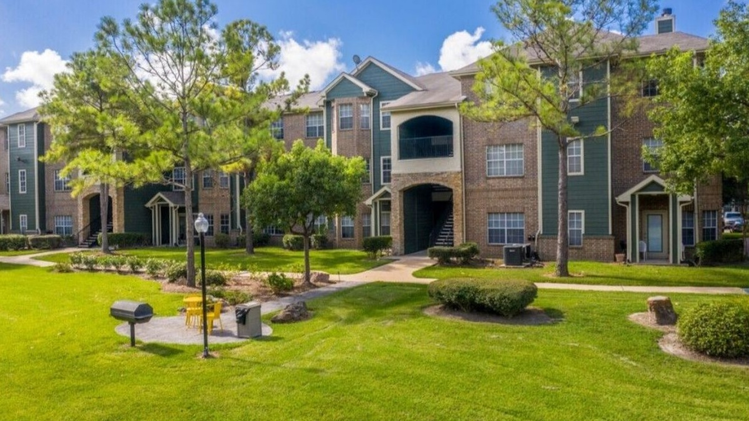 Charming apartment complex with beautiful brick and green accents, surrounded by lush greenery and vibrant trees, featuring an inviting outdoor seating area and peaceful pathways.