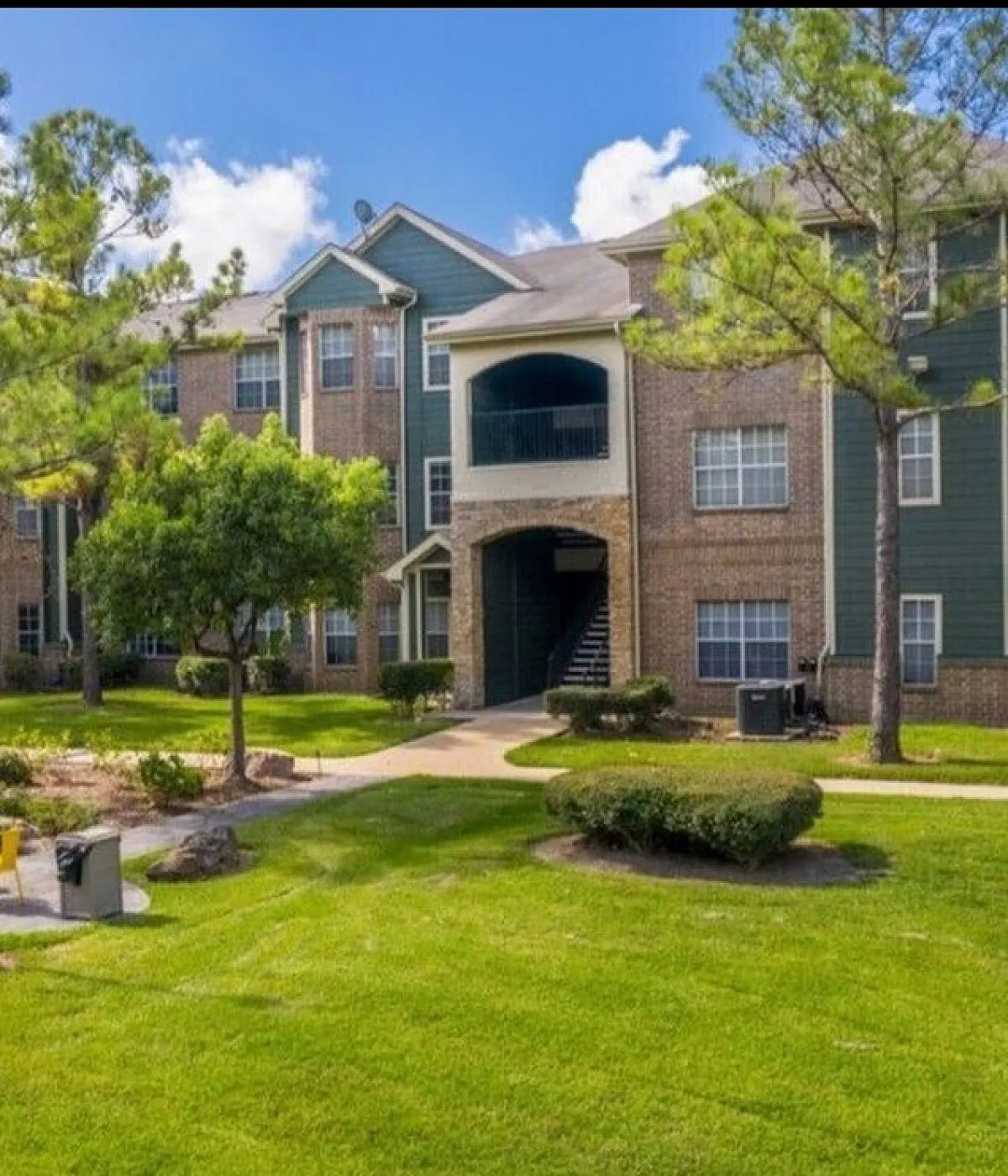 Charming apartment complex with lush green lawns, mature trees, and welcoming outdoor seating area, perfect for relaxation and community activities.
