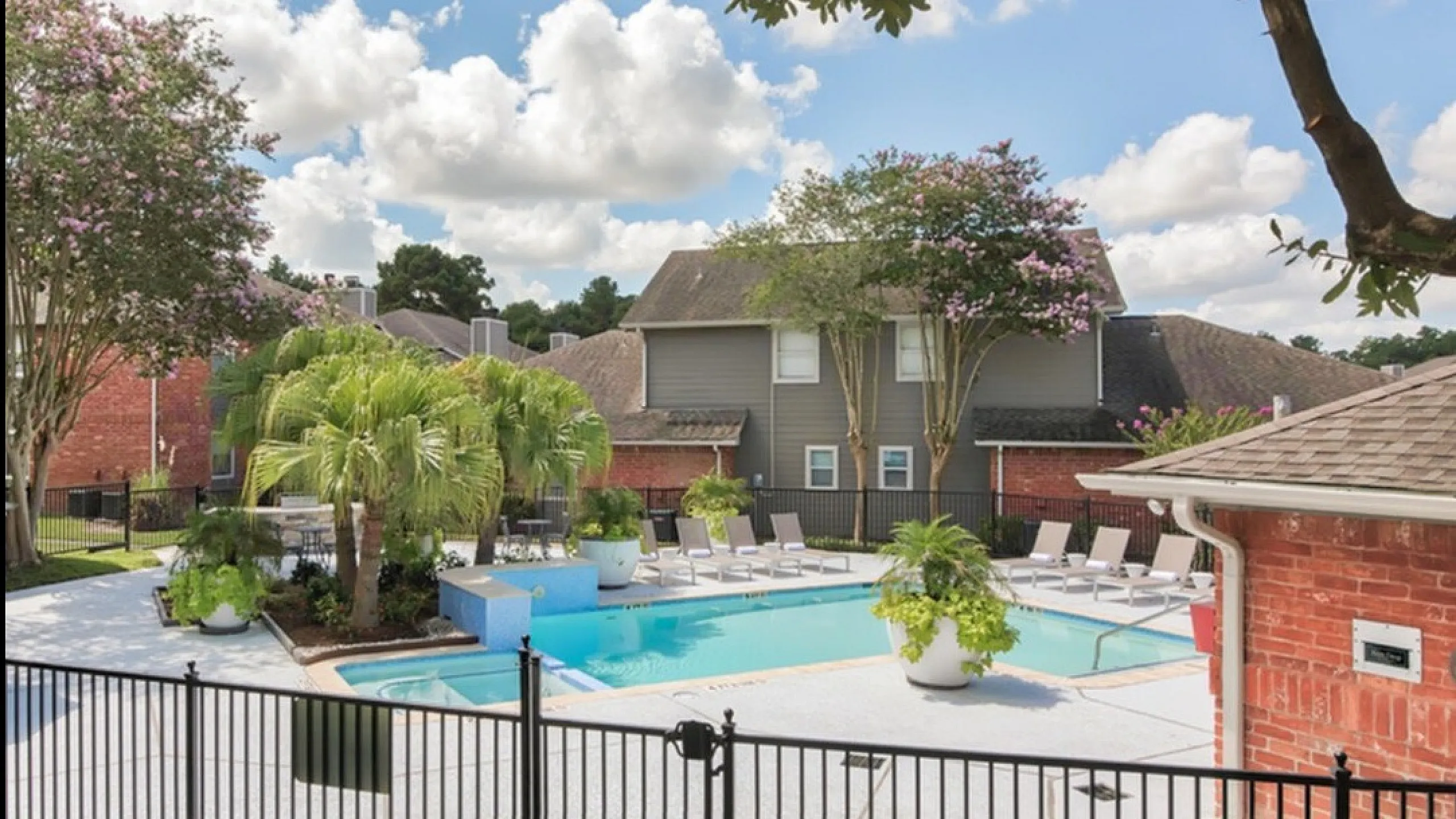 Charming apartment community with a sunlit pool area, surrounded by lush greenery and stylish lounge chairs.