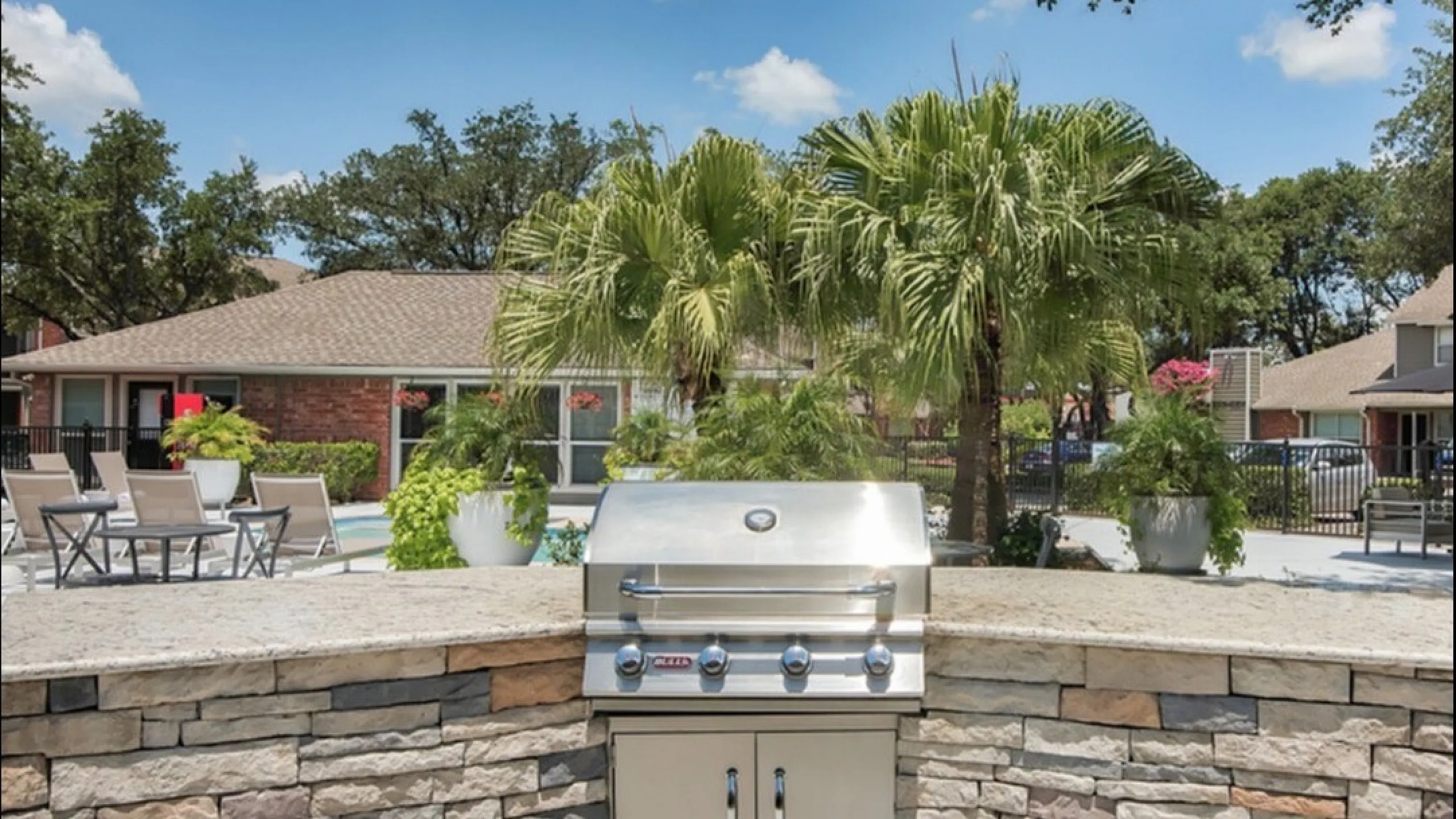 Luxurious outdoor area featuring a modern stone patio with a grill, surrounded by lush greenery and stylish seating under a clear blue sky.
