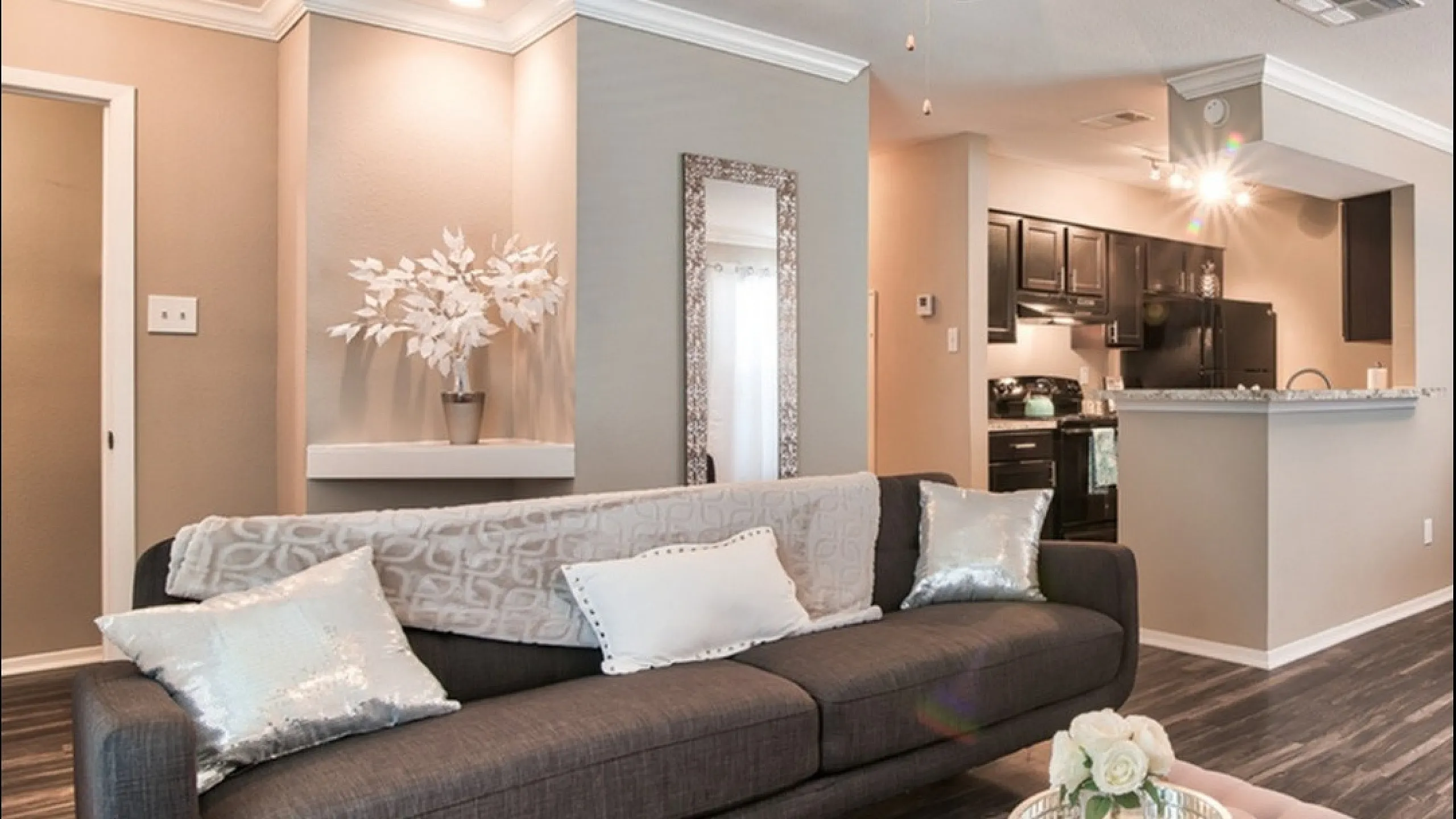 Elegant apartment living room with a plush gray sofa, decorative pillows, and modern ceiling fan, seamlessly connected to a sleek kitchen.