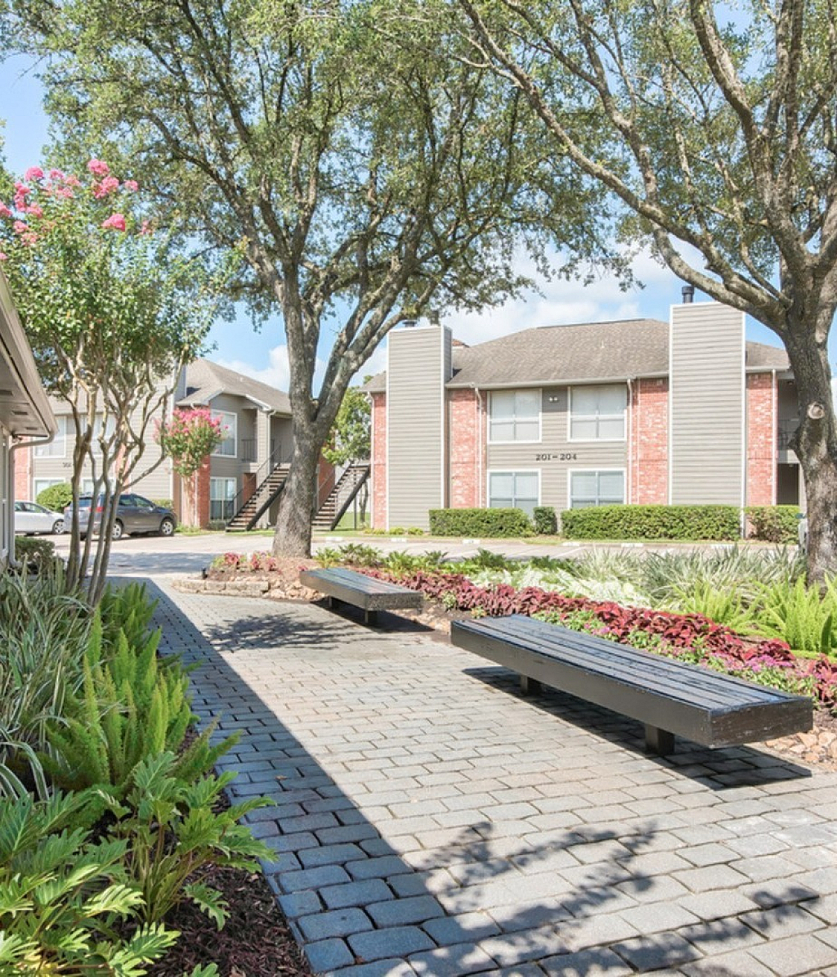 Charming apartment community with lush landscaping, brick exterior buildings, and inviting outdoor seating area under mature trees.