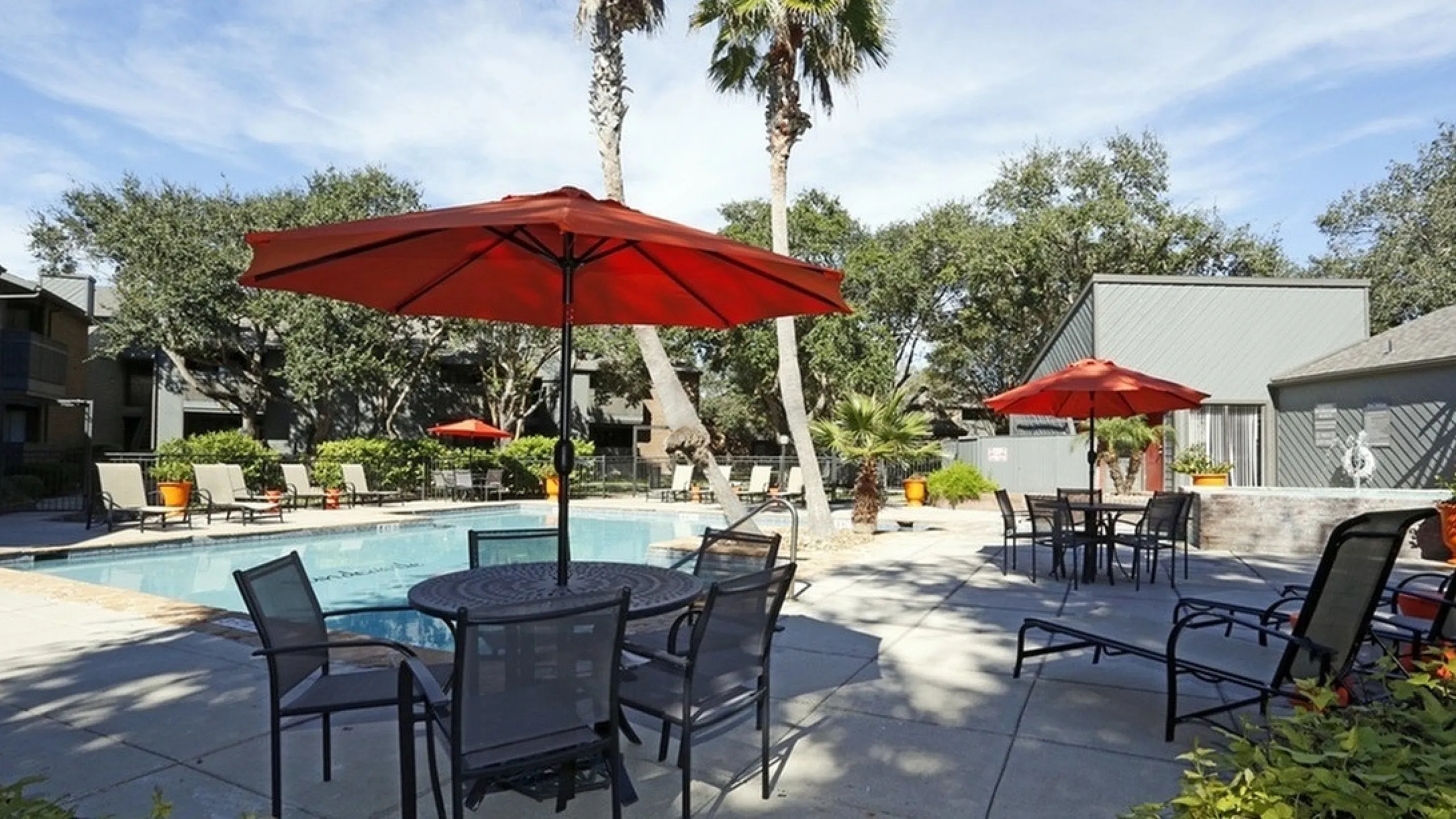 Inviting outdoor pool area with modern lounge chairs and vibrant red umbrellas surrounded by lush greenery and palm trees.