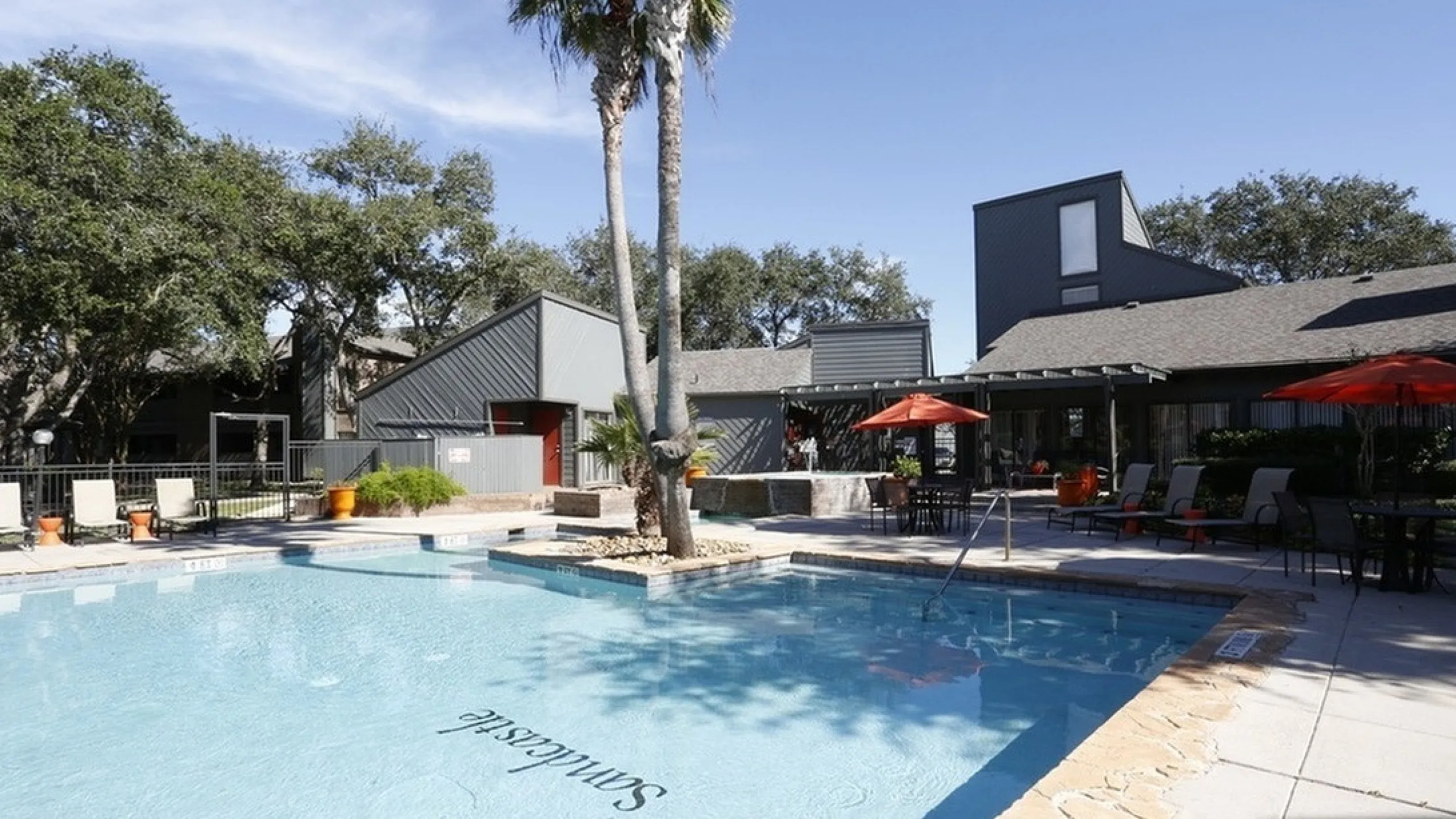 Sunny apartment complex featuring a sparkling swimming pool, shaded lounge chairs, and lush palm trees for a resort-style living experience.