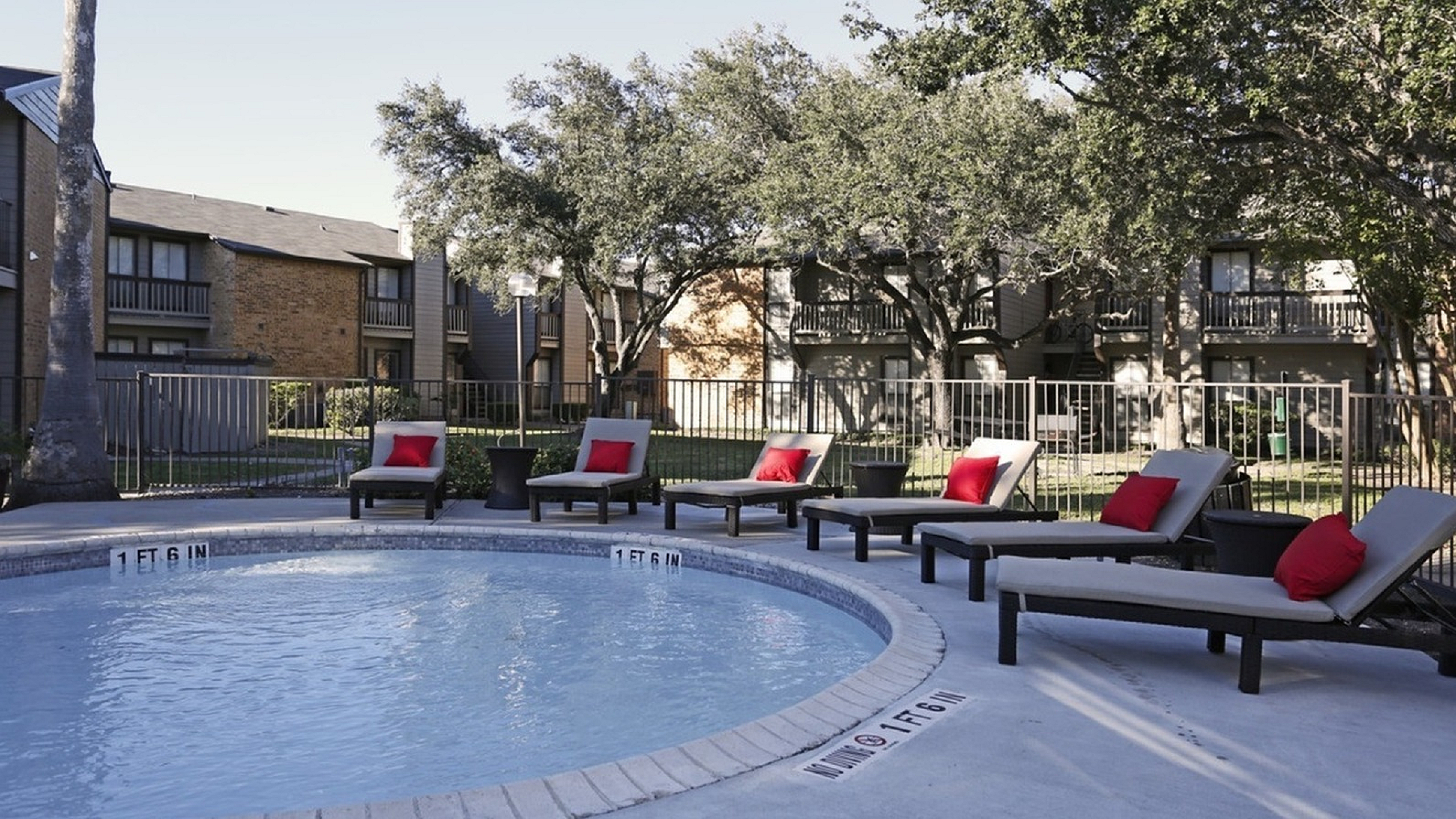 Inviting outdoor pool area with stylish lounge chairs and vibrant red cushions, surrounded by lush greenery and modern apartment buildings.