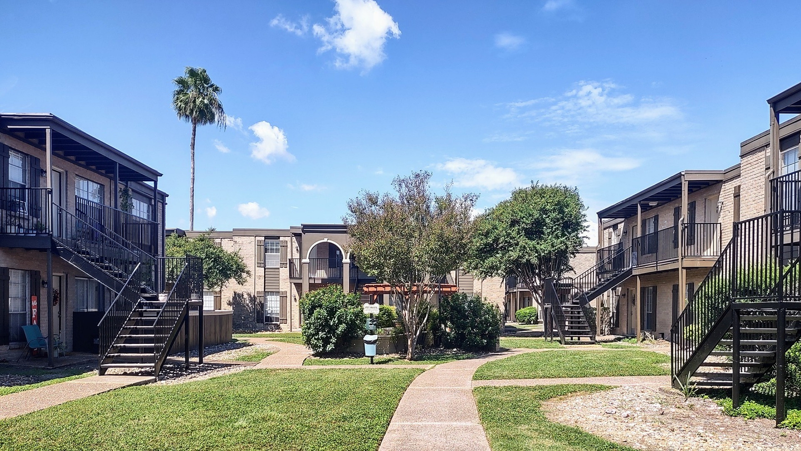 Charming apartment courtyard with lush greenery, modern staircases, and a serene atmosphere under a bright blue sky.