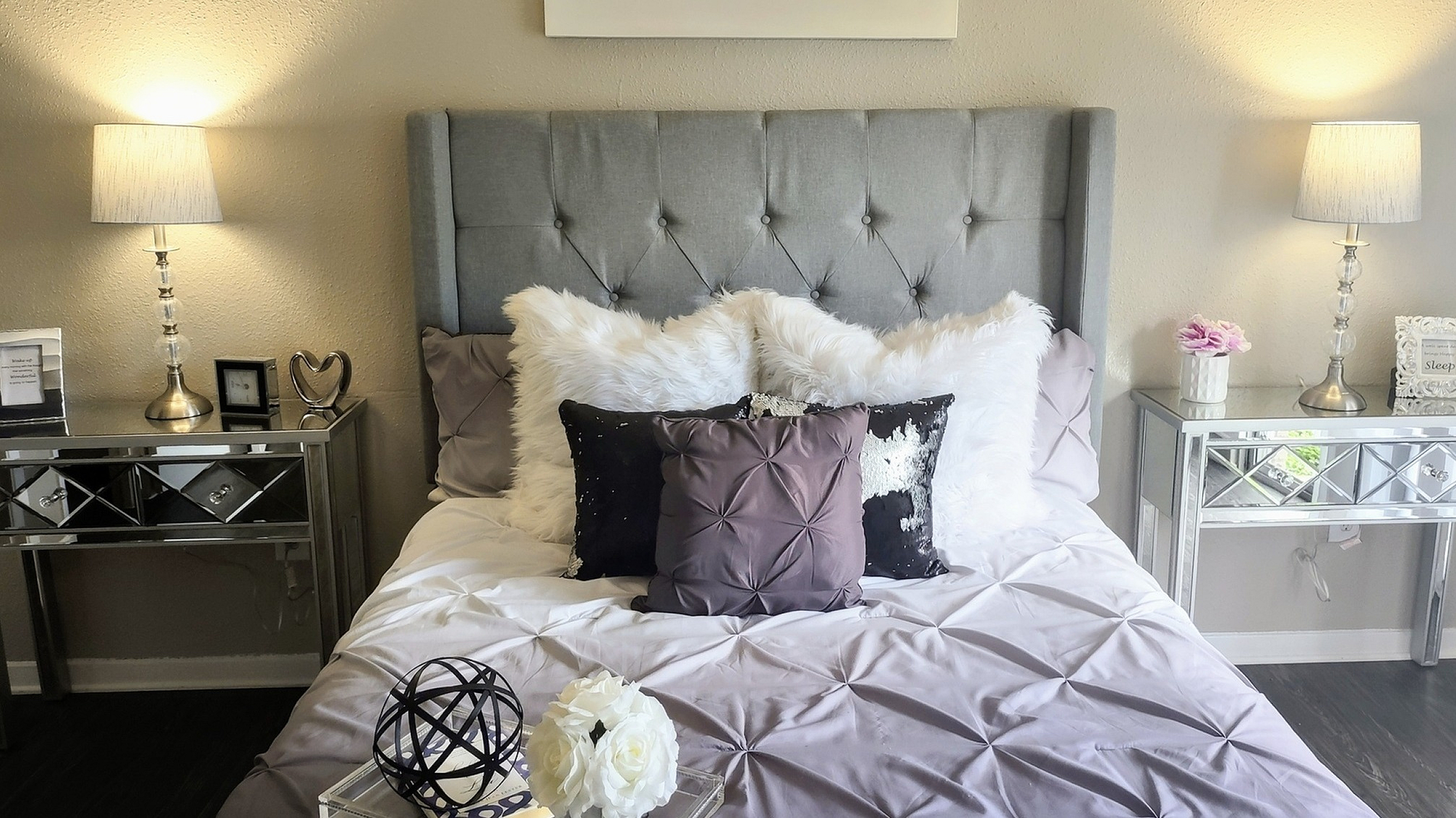 Elegant bedroom with a plush gray headboard, adorned with luxurious pillows, mirrored side tables, and chic decor, creating a serene and stylish ambiance.