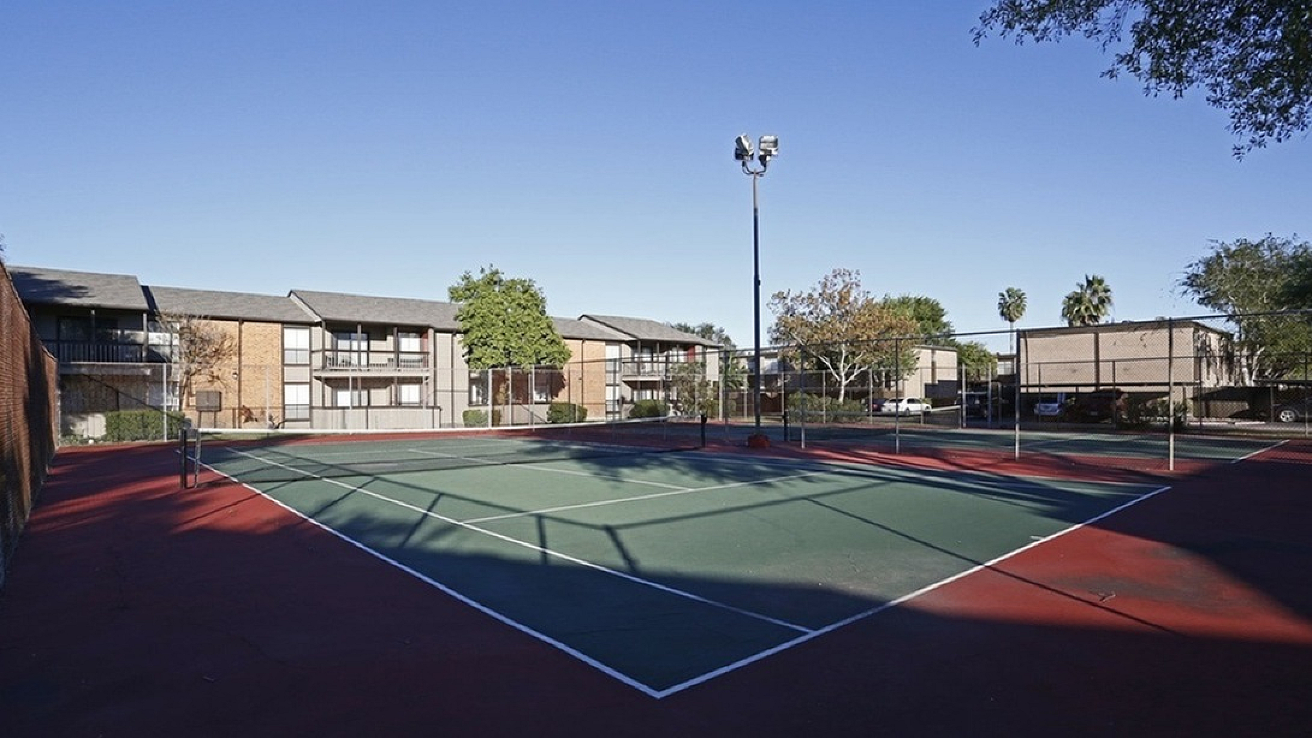 Sunny outdoor tennis court surrounded by modern apartment buildings and lush greenery.