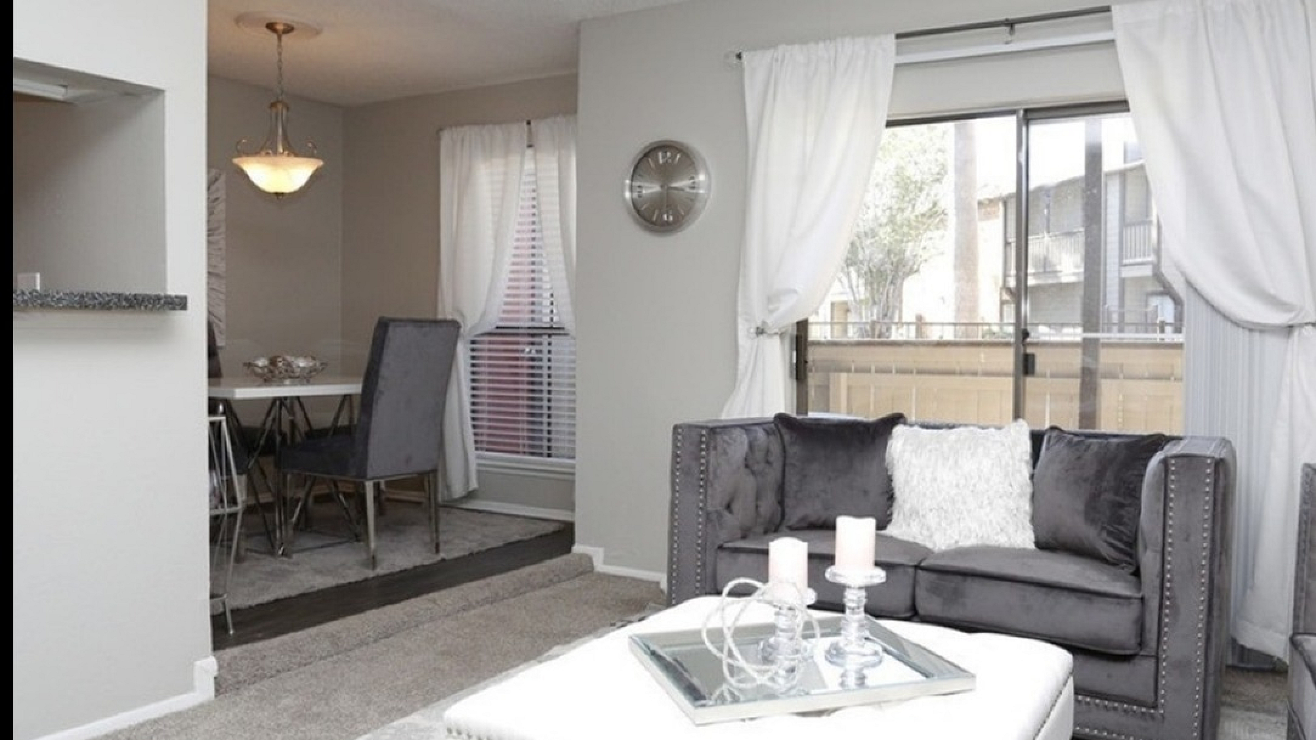 Modern living room with elegant gray sofa, plush white ottoman, and a stylish dining area. Natural light floods through large windows creating a bright, inviting atmosphere.