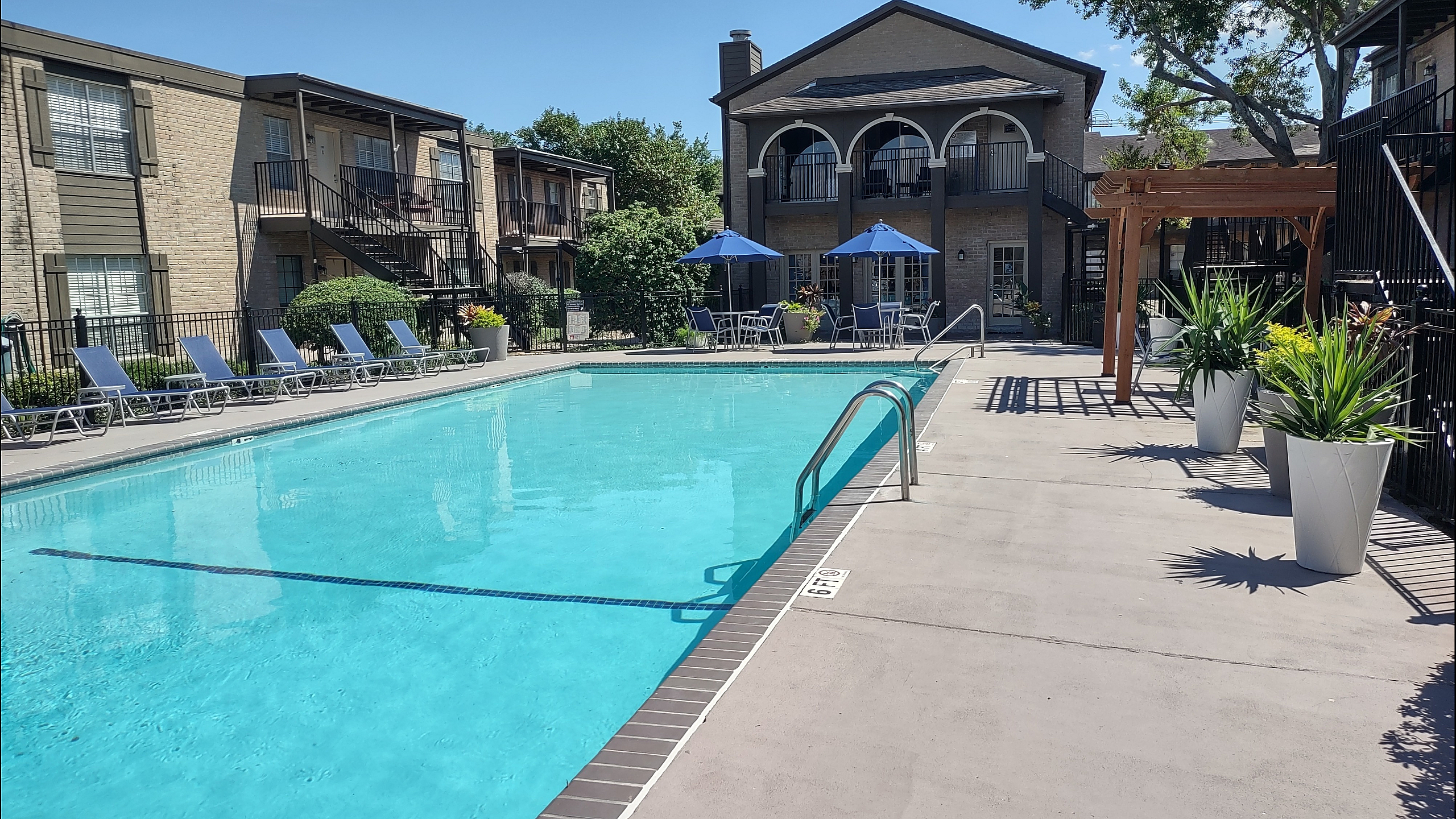 Sunny apartment complex with a sparkling pool, surrounded by elegant patio furnishings and lush greenery, offering a perfect blend of relaxation and style.